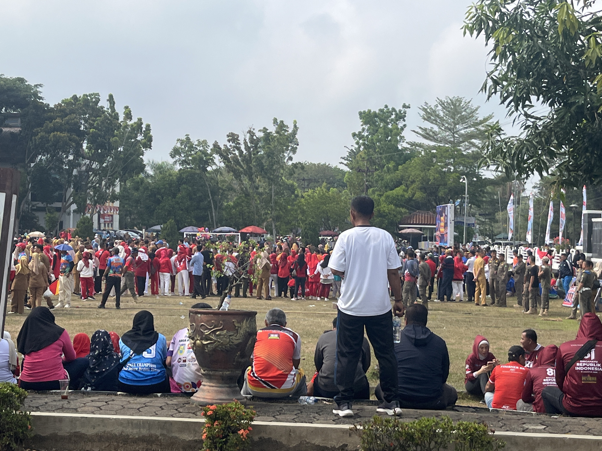 This is an outdoor scene at what appears to be a public gathering or event in Sekayu, Indonesia. The foreground shows several individuals seated on the ground, some facing away from the camera. They are wearing casual clothing, including headscarves and t-shirts. A large, ornate pot is also in the foreground.
The background is filled with a crowd of people, many wearing red and white attire, suggesting a patriotic or celebratory occasion. There are also individuals in what appear to be uniforms, possibly security personnel or event staff. Tents and flags are visible in the distance, along with trees and buildings. The overall atmosphere seems to be one of a community event or festival.