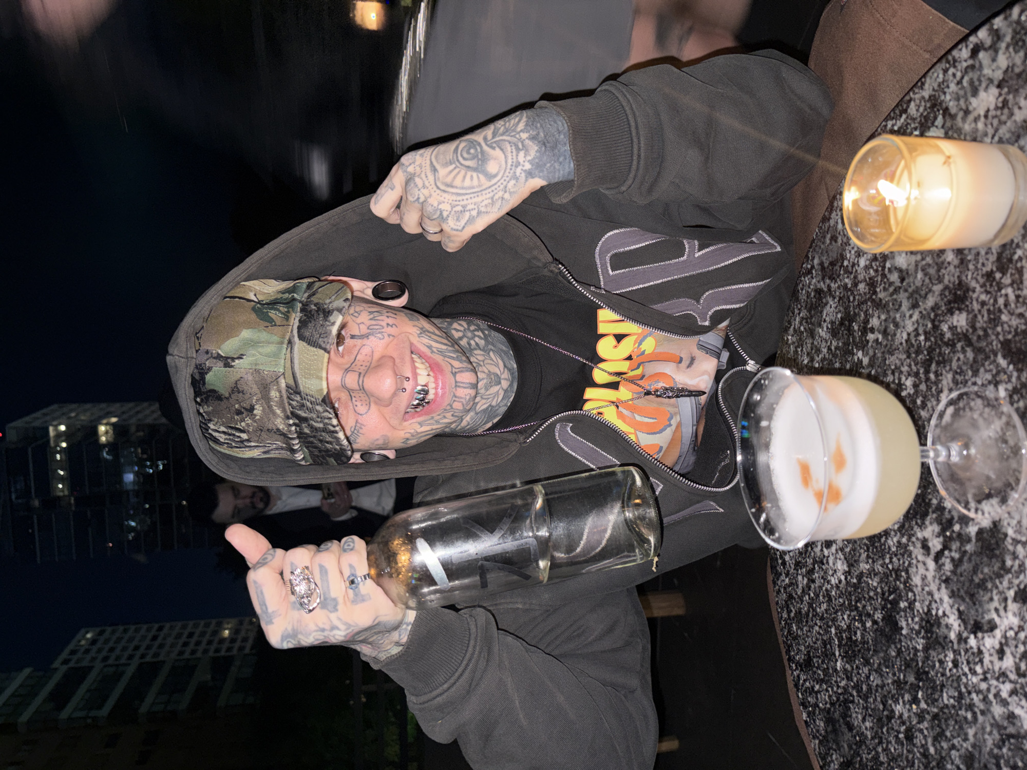 A heavily tattooed person wearing a camouflage cap and a dark hooded sweatshirt is shown at a table with a cocktail and a candle. The person is holding an empty glass bottle. Another person is partially visible behind them. The background shows a night scene with buildings. The location is the city of Buenos Aires, Argentina.