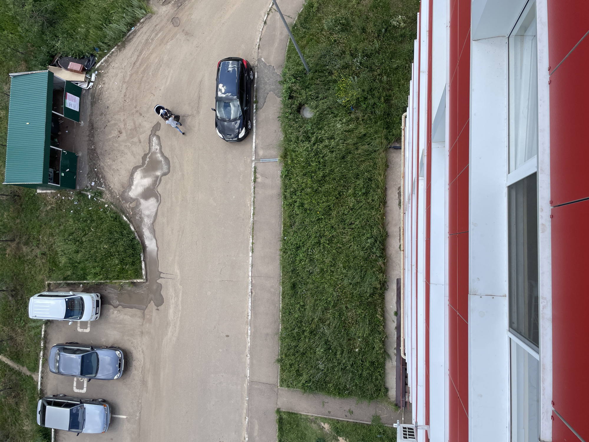 An aerial view shows a parking lot with several cars, a green recycling station, and a person pushing a stroller. A black car is parked on the asphalt, and a white van, a blue car, and a silver car are parked further up. A person is pushing a stroller near the recycling station. The recycling station is a green metal structure with open bays, surrounded by debris. Beyond the parking lot is a patch of green grass. In the foreground, the red and white exterior of a building is visible, suggesting the image was taken from a high vantage point. The scene appears to be in a residential area of Laishevo, Russia, given the presence of apartment buildings and common urban infrastructure.