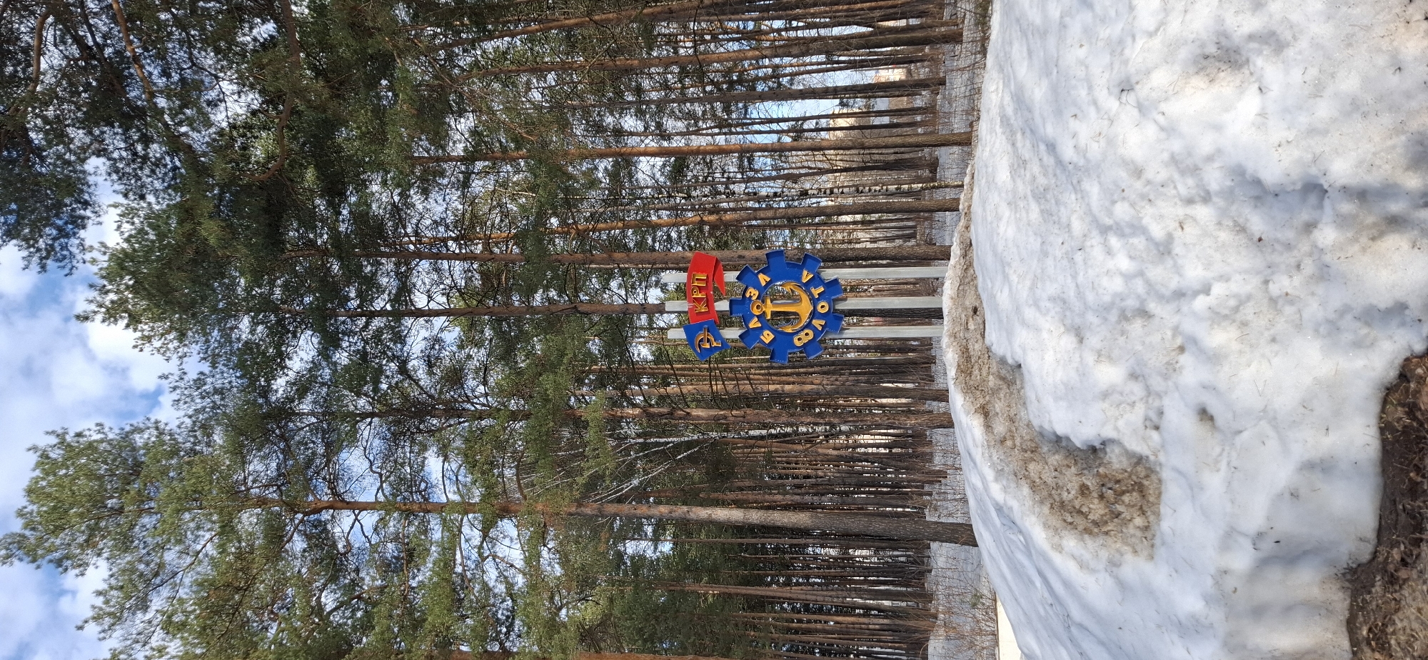 The image depicts an outdoor scene in Chaikovsky, Russia, likely during winter or early spring, characterized by snow cover and pine trees under a partially cloudy sky. In the midground, a prominent, colorful informational sign is erected on three vertical white posts. This sign features a large blue gear-shaped emblem with a gold anchor at its center. Gold Cyrillic text, "ЧАЙКОВСКОГО" (Chaikovsky), is inscribed on the upper curve of the gear, and "БАЗОВОЙ ФЛОТА" (Base Fleet) is on the lower curve. Above the main emblem, two smaller flags are positioned: one on the left is blue with a red "РКРП" (RKRP, Russian Communist Workers' Party) emblem, and one on the right is red with white Cyrillic text "КПП" (KPP, meaning Checkpoint).
The foreground consists of an uneven snow-covered ground, featuring patches of dirty, brown-tinged snow and exposed earth, suggesting melting or debris. A track or depression of dirty snow is visible, extending from the lower center towards the midground. The background is dominated by a dense, vertical forest of tall, slender pine trees with some deciduous trees scattered among them, all under a bright blue sky with scattered white clouds. No individuals or animals are present in the scene.