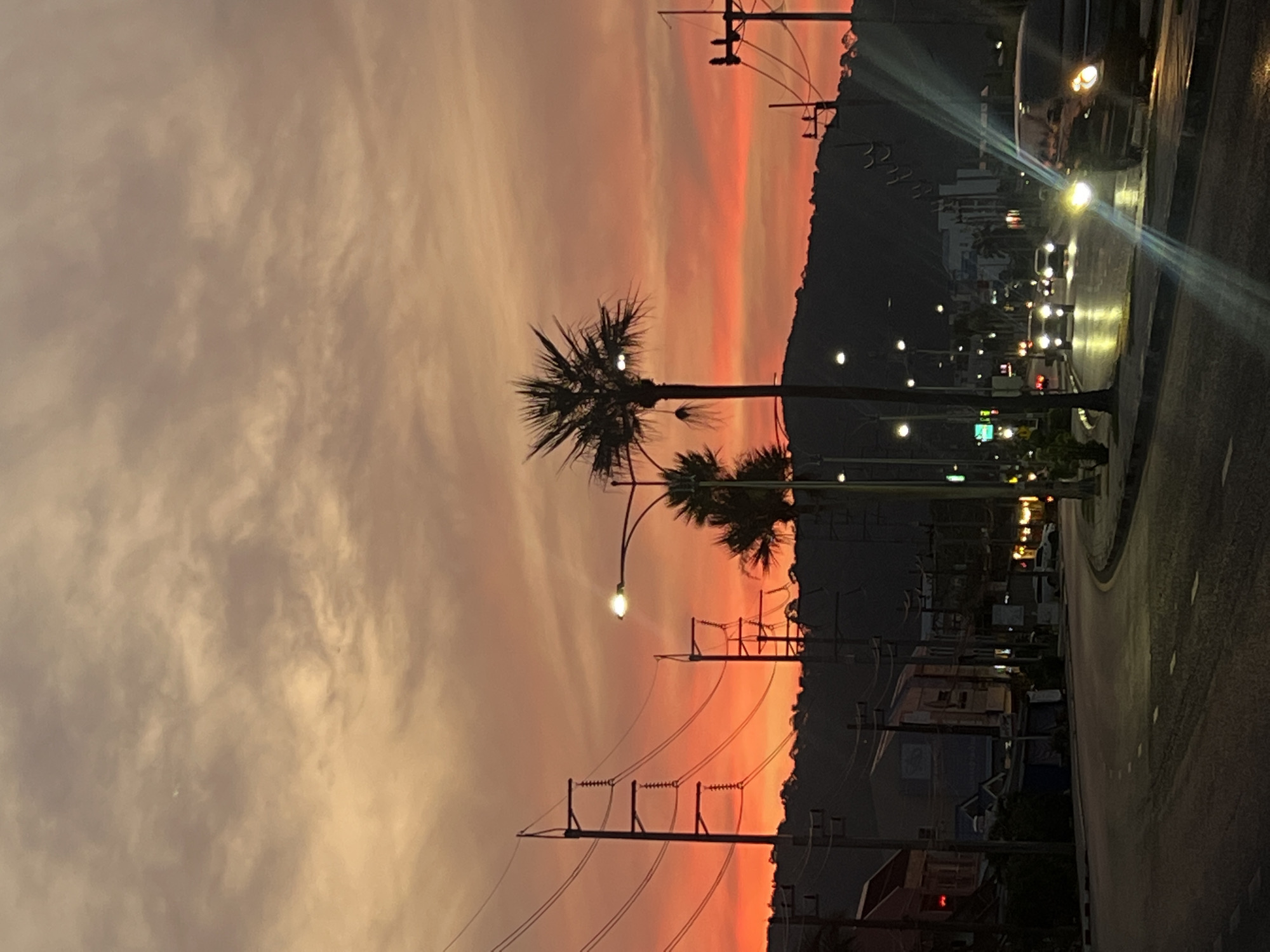 The image captures a scene at dusk in Phuket City Municipality, Thailand. A palm tree stands prominently against a backdrop of orange and pink hues in the sky. Utility poles with electrical wires are scattered across the landscape. A road runs along the right side of the frame with street lights illuminating the path and vehicles with headlights on. The background includes buildings and a silhouette of a hill.