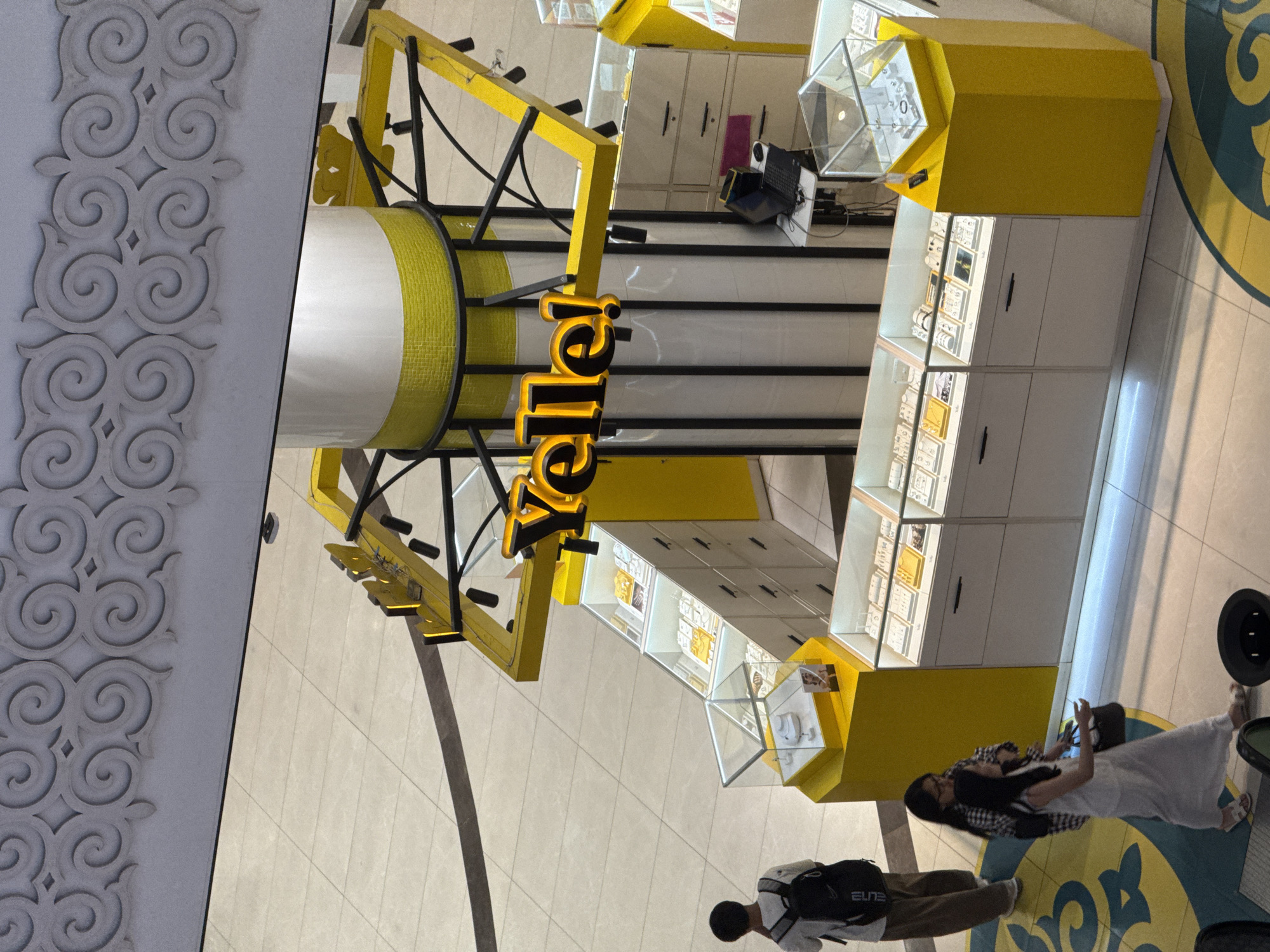 In an indoor shopping mall, a bright yellow jewelry kiosk, branded "Yelle!", is the central focus. The kiosk is hexagonal and features multiple glass display cases showcasing various jewelry items. A white cylindrical pillar, adorned with a yellow mosaic band, stands at the kiosk's center. Overhead, a decorative grey frieze with swirling patterns adorns the upper wall. On the tiled floor, a large yellow and teal circular motif is visible. Three individuals are present: a young person with a backpack stands to the left, facing away from the camera. Two women are walking from the left to the right in front of the kiosk, one carrying a dark handbag. The environment appears clean and well-lit, typical of a commercial space.