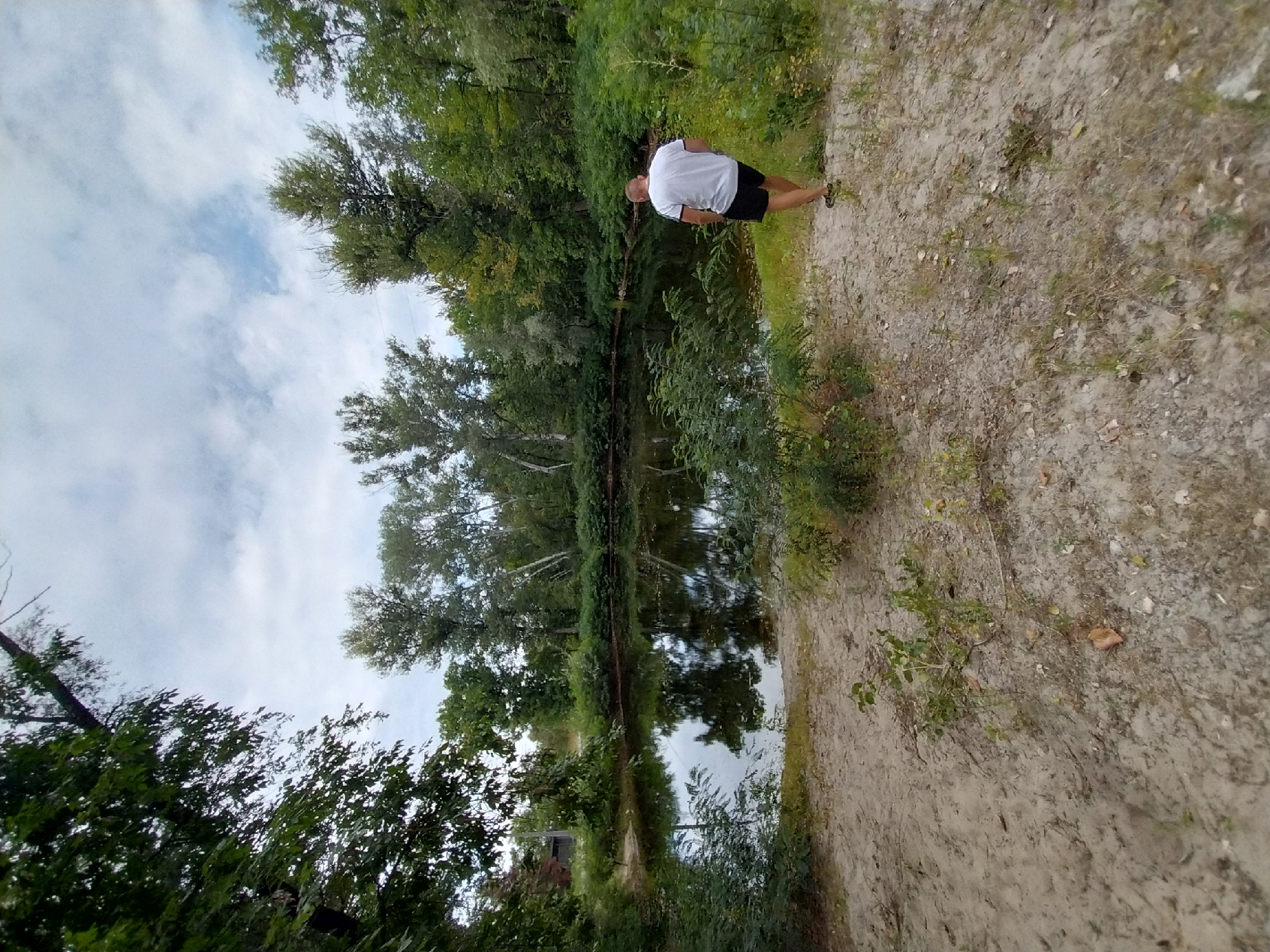 A man in a white t-shirt and black shorts walks away from the camera towards a body of water. The scene is set in a natural environment with trees and foliage surrounding the water. The ground in the foreground is sandy with sparse vegetation. The sky above is partly cloudy.