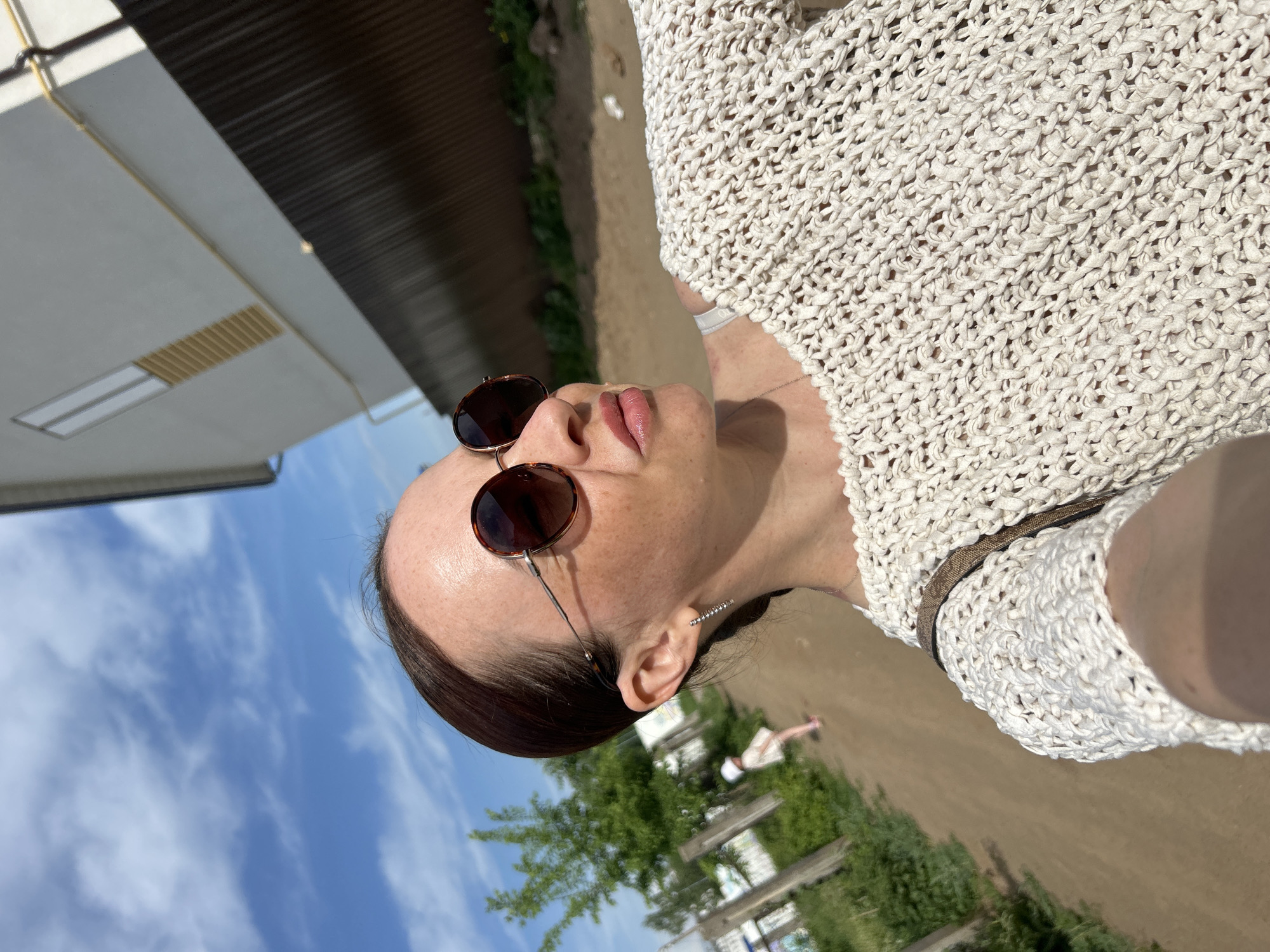 A woman wearing round sunglasses and a white knit top is pictured in an outdoor setting. The sky is visible with scattered clouds. A building with a brown corrugated roof is in the background. There is vegetation, including trees and greenery, present. A fence and a person in white attire are also visible in the distance. The image was likely taken in Chaikovsky, Russia.