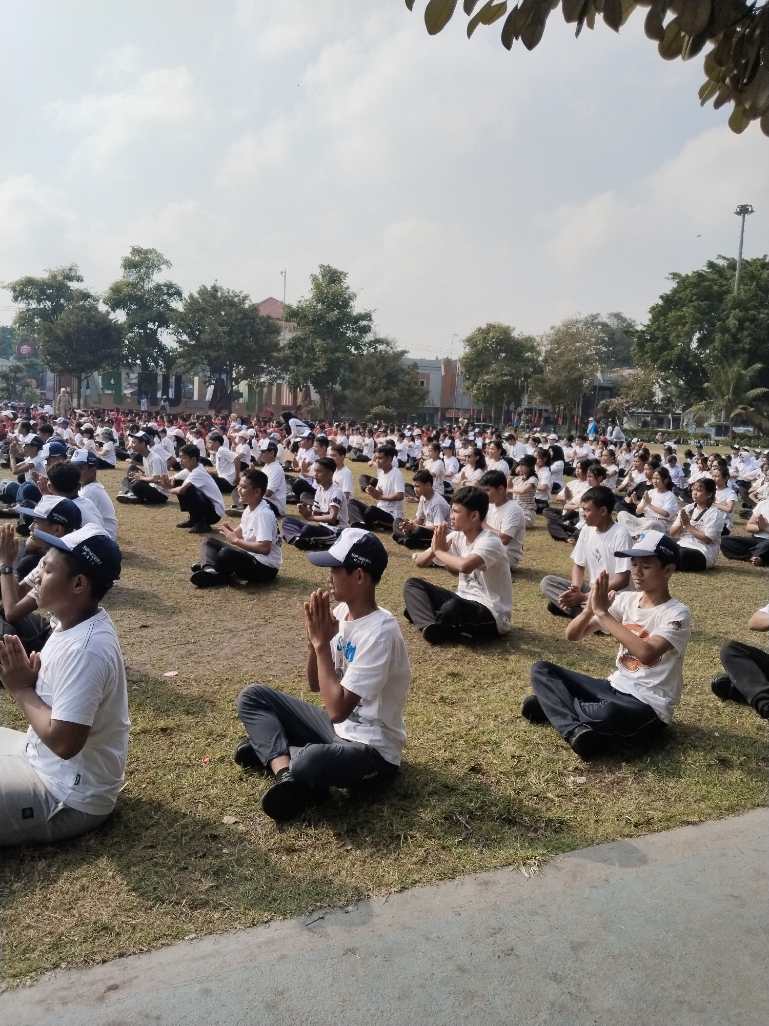 A large group of people, likely students, are seated on a grassy field performing a group activity that appears to be meditation or a similar exercise. They are all wearing white t-shirts and many are wearing white baseball caps. The participants are arranged in rows and columns, facing forward. The environment is outdoors with trees and buildings visible in the background. The sky is cloudy. The activity seems to be taking place in an open public space, possibly a school field or park, within Kecamatan Pati, Indonesia.