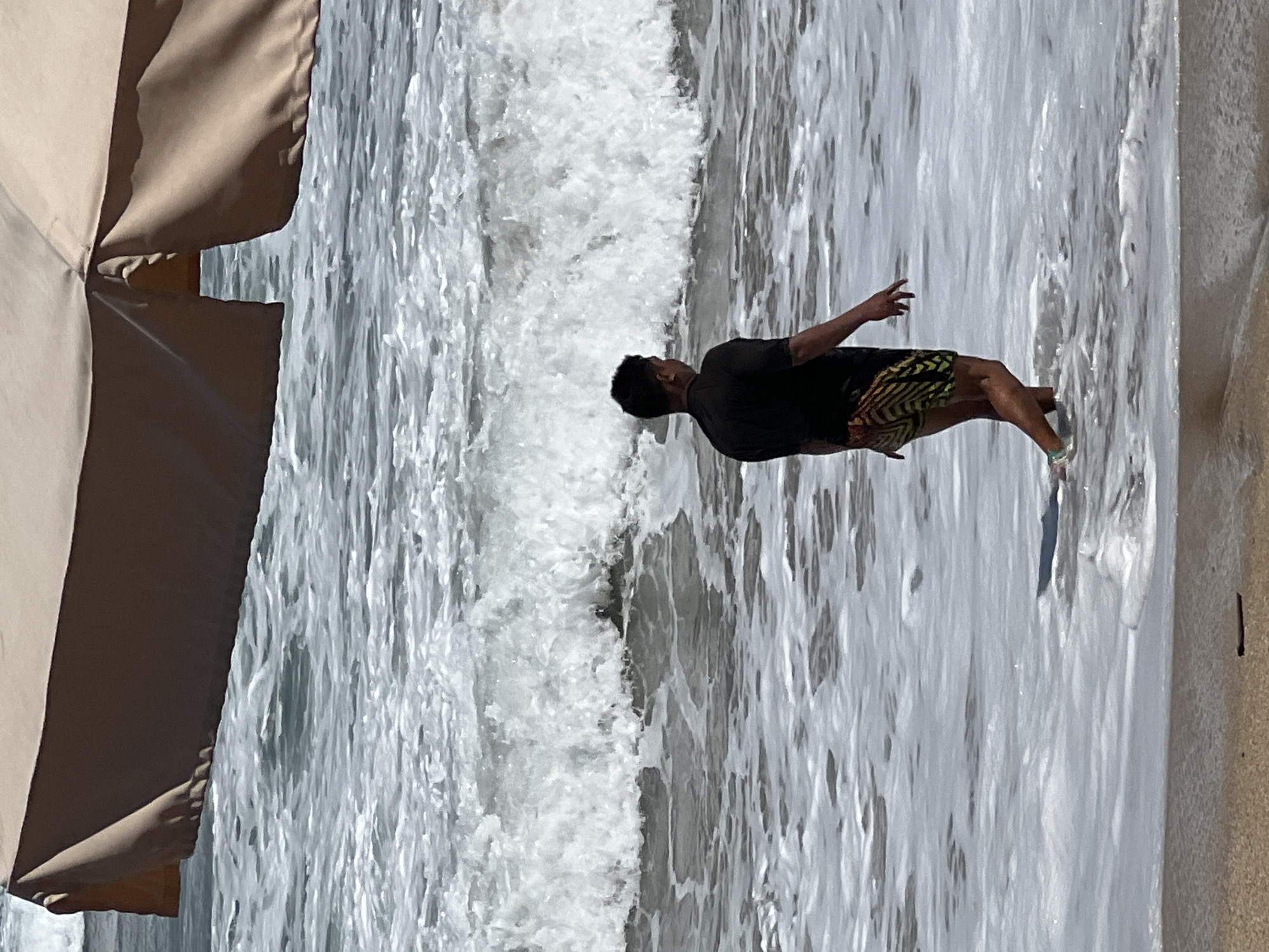 A man walks into the ocean at Kuta Beach, Bali, Indonesia. The man is facing away from the camera, wearing a black t-shirt and patterned swim trunks. He is knee-deep in the water, with waves breaking around him. In the foreground, a beach umbrella casts a shadow over the sand and sea. The ocean is choppy with white foam.