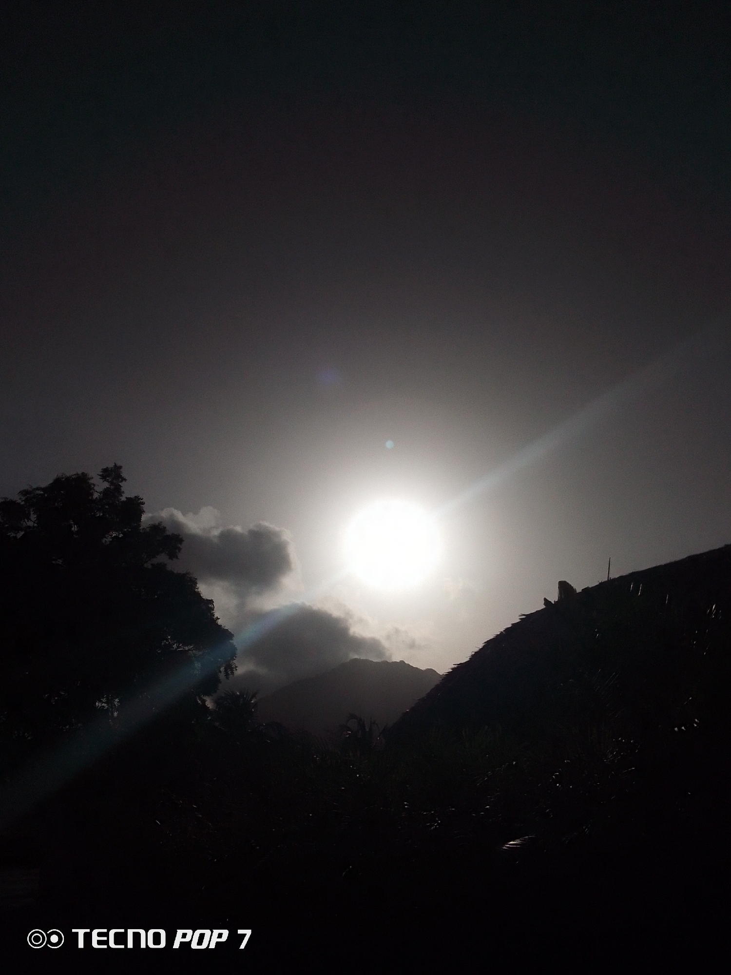 The image shows a landscape scene in Pedro González, Venezuela, under what appears to be a bright sun partially obscured by clouds. A dark silhouette of foliage and a thatched roof are visible in the foreground. In the background, there are silhouettes of mountains or hills. The overall scene has low contrast.