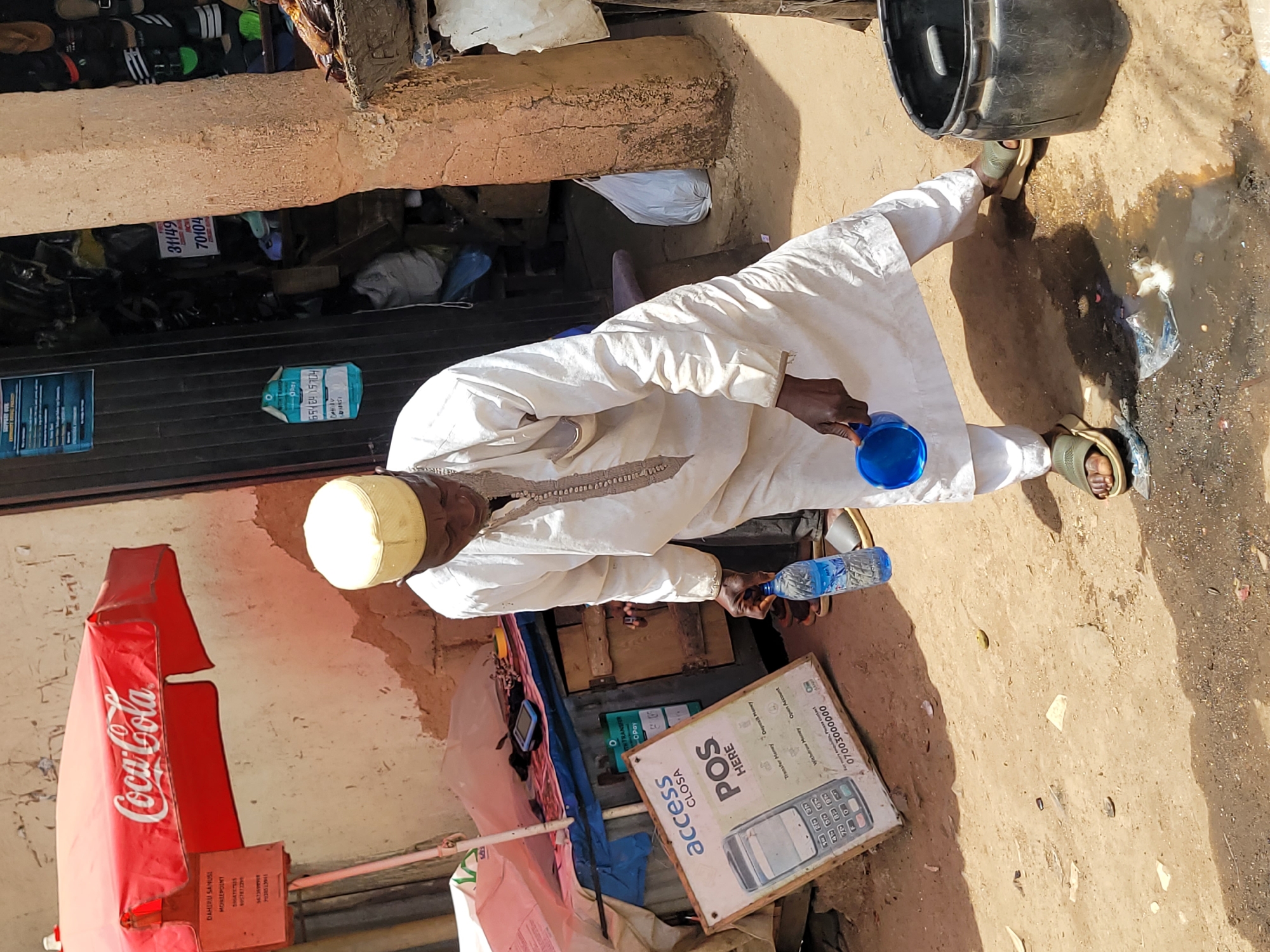 A man dressed in white stands in a dusty environment in what appears to be a commercial area of Jos, Nigeria. He is wearing a white robe and a yellow cap, holding a blue plastic cup in his right hand and a bottle of water in his left. He is standing next to a muddy puddle with some refuse floating in it.
Behind the man is a small business setup. There is a Coca-Cola branded umbrella, a POS (Point of Sale) sign for Access CLOSA (a banking agent service), and what appears to be a small wooden stall with items displayed. The stall is covered by a dark blind. The POS sign advertises various banking services such as money transfers, deposits, withdrawals, and account openings, with a phone number listed. Numerical markings are visible on another sticker.
The ground is sandy and littered with debris. A black bucket is positioned on the right of the scene, with the wall of a building on the left. The overall impression is of a typical small-scale commerce scene in an urban Nigerian setting.
