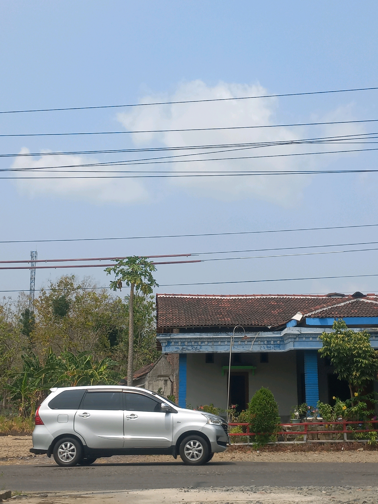 The image captures a street scene in Baturetno, Indonesia, on a clear day with a bright blue sky dotted with white clouds. Multiple power lines stretch horizontally across the upper portion of the frame. In the foreground, a silver minivan is parked or driving along a paved road. To the right of the vehicle, a house with a red tiled roof and blue accents is visible. The house features a covered porch area with blue pillars. Behind the house, various trees and vegetation can be seen, including a tall tree with a thin trunk. A telecommunications tower is also present in the distance. The overall environment appears to be a mix of residential and natural elements.
