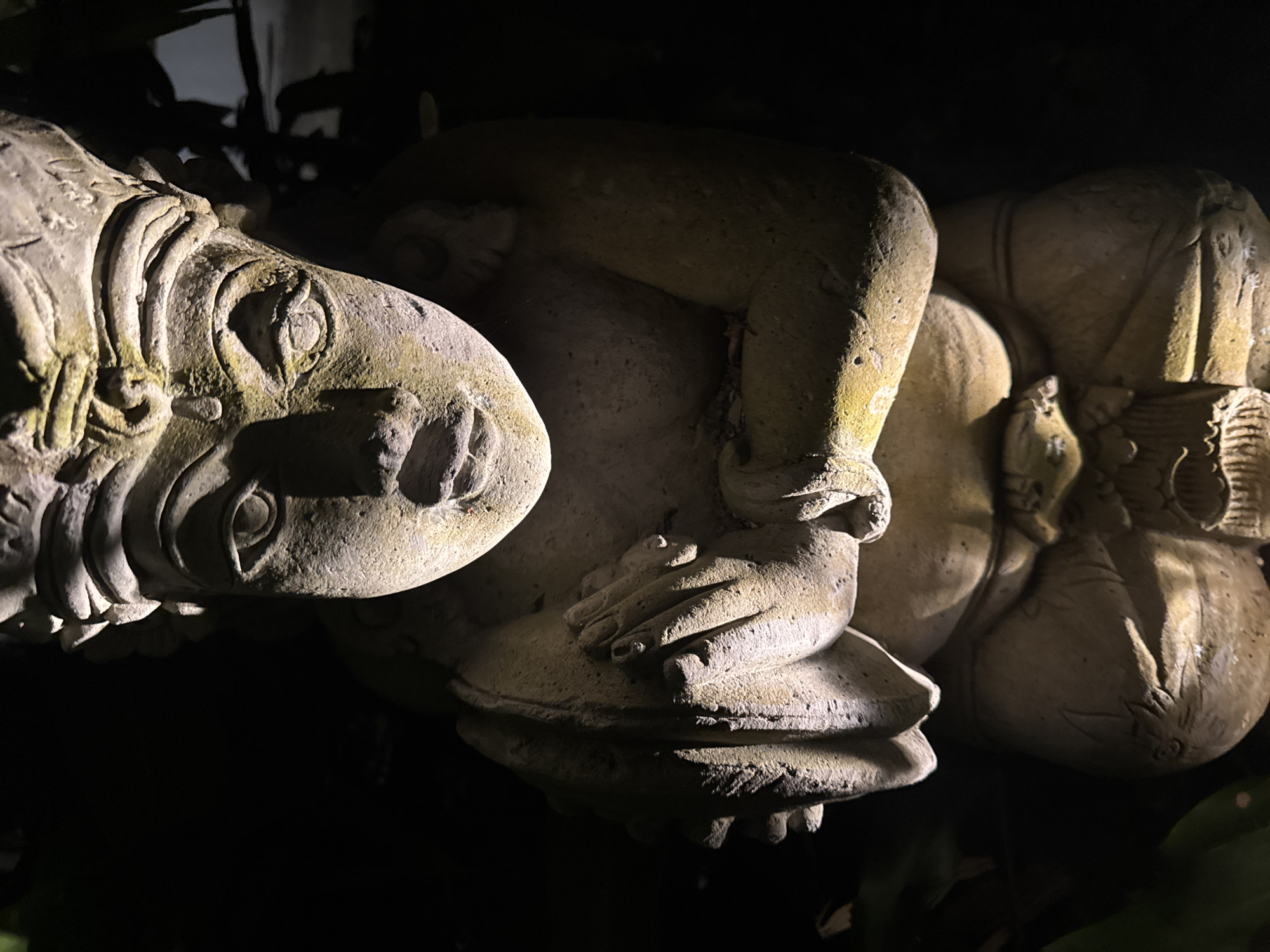 The image shows a close-up of three weathered, stone-like statues bathed in dim, directional light against a dark, undefined background. The leftmost statue depicts a stylized animal head with intricate patterns and a prominent eye. Adjacent to it is a human-like figure with its arm and hand resting on the statue below. The third statue is partially visible on the right, seemingly another rounded figure, perhaps part of the main statue. The environment is obscured in darkness, suggesting an outdoor setting at night. The statues appear to be part of a larger installation or garden feature in Kuta Selatan, Indonesia.