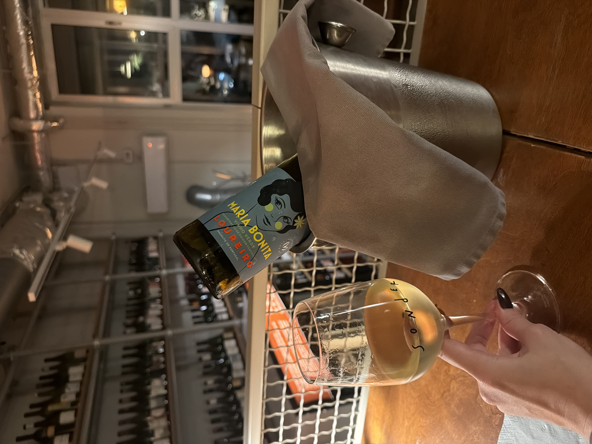 The image depicts an indoor scene at Sonder Wines, located in Skadarlija, Belgrade, Serbia. A fair-skinned hand, with dark nail polish on the thumb and index finger, holds a clear stemmed wine glass containing a pale yellow liquid. The word "Sonder" is visible on the glass. To the right, a silver-colored ice bucket, partially covered by a light brown fabric napkin, holds a green glass wine bottle. The bottle's label features a stylized face and the prominent text "MARIA BONITA" and "LOUREIRO VINHO VERDE". The immediate surface is a light brown wooden table with a visible seam. In the background, multiple shelves are stacked with numerous wine bottles, indicating a wine shop or bar setting. To the right, a window reveals blurred outdoor lights, suggesting an evening scene. An air conditioning unit is mounted on the wall, and exposed ductwork is visible on the ceiling. A white metal grid structure is present in the mid-background. The lighting is dim throughout the scene.