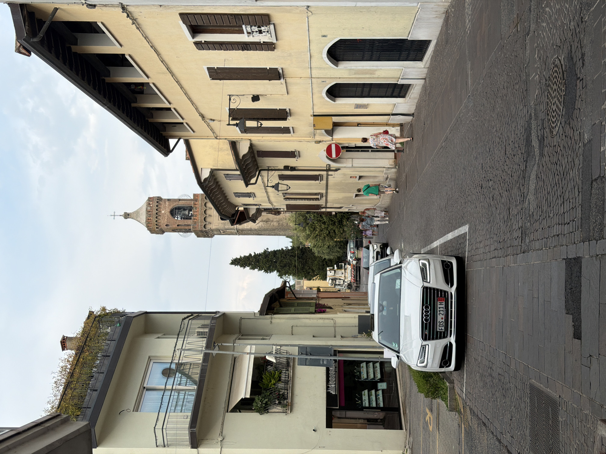 A white Audi A8 sedan is parked on a cobblestone street in front of a building. The car has a license plate that reads "FDS 211 H". The street is lined with buildings, one of which has a balcony with plants. In the background, a church tower is visible. Several people are walking on the street. The sky is overcast. The image appears to have been taken in the town of Padenghe sul Garda, Italy.