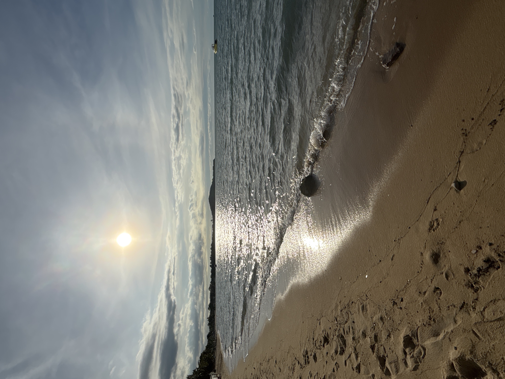 The image shows a beach in Baan Mae Nam, Thailand. The scene depicts a shoreline with light brown sand meeting the calm, gray-blue water of the sea. The sun is positioned high in the sky, filtering through a layer of thin clouds, creating a bright, diffused light. The reflection of the sun glitters across the surface of the water towards the shoreline. Small waves gently lap at the edge of the beach. Footprints are visible in the sand, suggesting recent activity. In the distance, there is vegetation lining the coast, and a small boat is on the water. A few rocks or dark patches are also visible on the beach near the water's edge. The image is oriented vertically.
