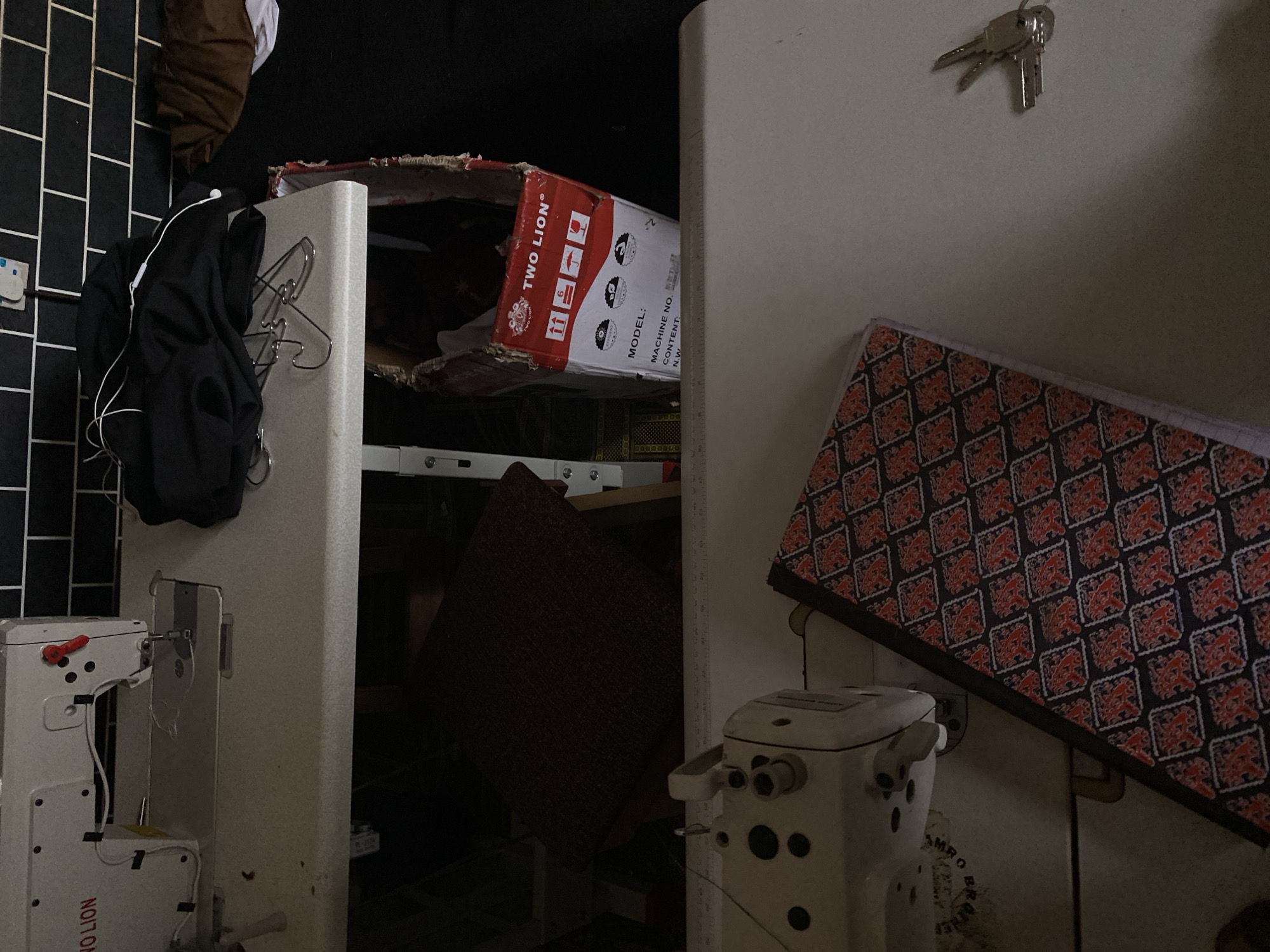 The image depicts a dimly lit room, possibly a workshop or studio, in Jalingo, Nigeria. On a workbench, a white sewing machine with the branding "TWO LION" is partially visible, along with its metal plate and needle. Next to it, a dark brown fabric is draped over another workbench. Below this, a cardboard box, also branded "TWO LION," lies on its side. It contains what appears to be part of a machine, with labels indicating "MODEL," "MACHINE NO.," and "CONTENT." A portion of a black garment with white earbuds is piled on the upper workbench, near several metal clothes hangers. In the foreground, another white sewing machine is visible, and a notebook with a red and black geometric pattern lies open on a surface. To the right, a small set of keys is placed on the surface. The background is a tiled wall with a white electrical outlet.