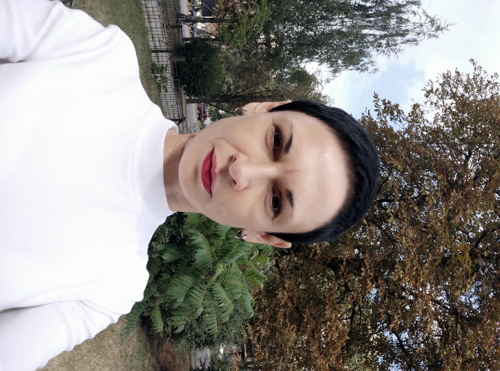 An outdoor medium shot shows a woman with short black hair and dark eyebrows, looking directly at the camera. She is wearing a white turtleneck shirt. Her lips are painted red. She has a silver hoop earring in her left ear. She is positioned in front of a blurred background of trees and a street. The trees on the right have brown leaves, and the trees on the left have green leaves. A metal fence is visible in front of the trees. A cloudy sky is overhead. The woman's facial expression is neutral. The overall scene suggests an urban environment in autumn. The location is Kyiv, Ukraine.