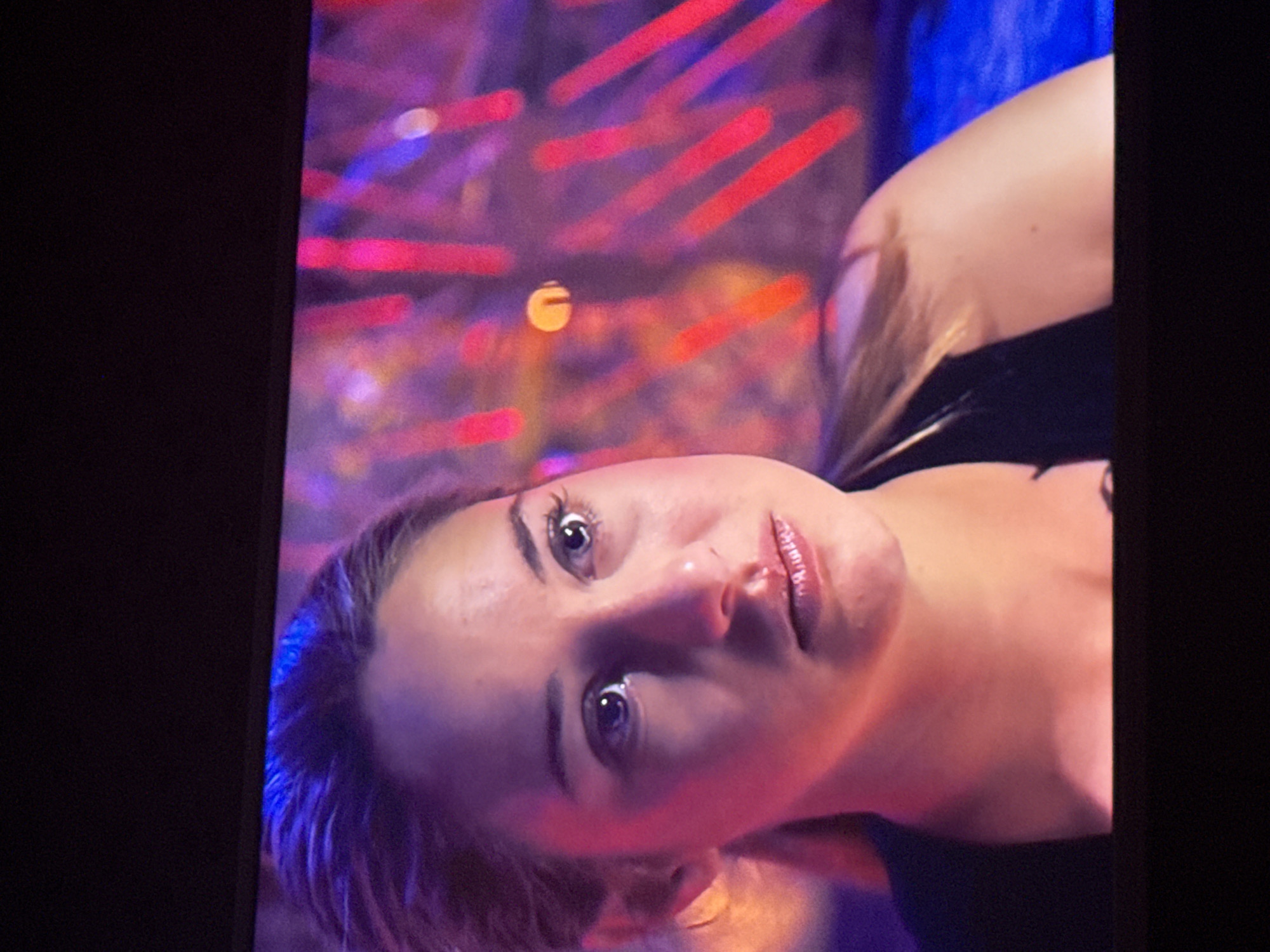 A close-up, slightly angled shot of a woman's face is displayed on a television screen, set against a blurred background of red and blue lights. The woman has fair skin, light-colored eyes, and glossy lips. She is wearing a dark garment. The setting and location are not specified within the image.