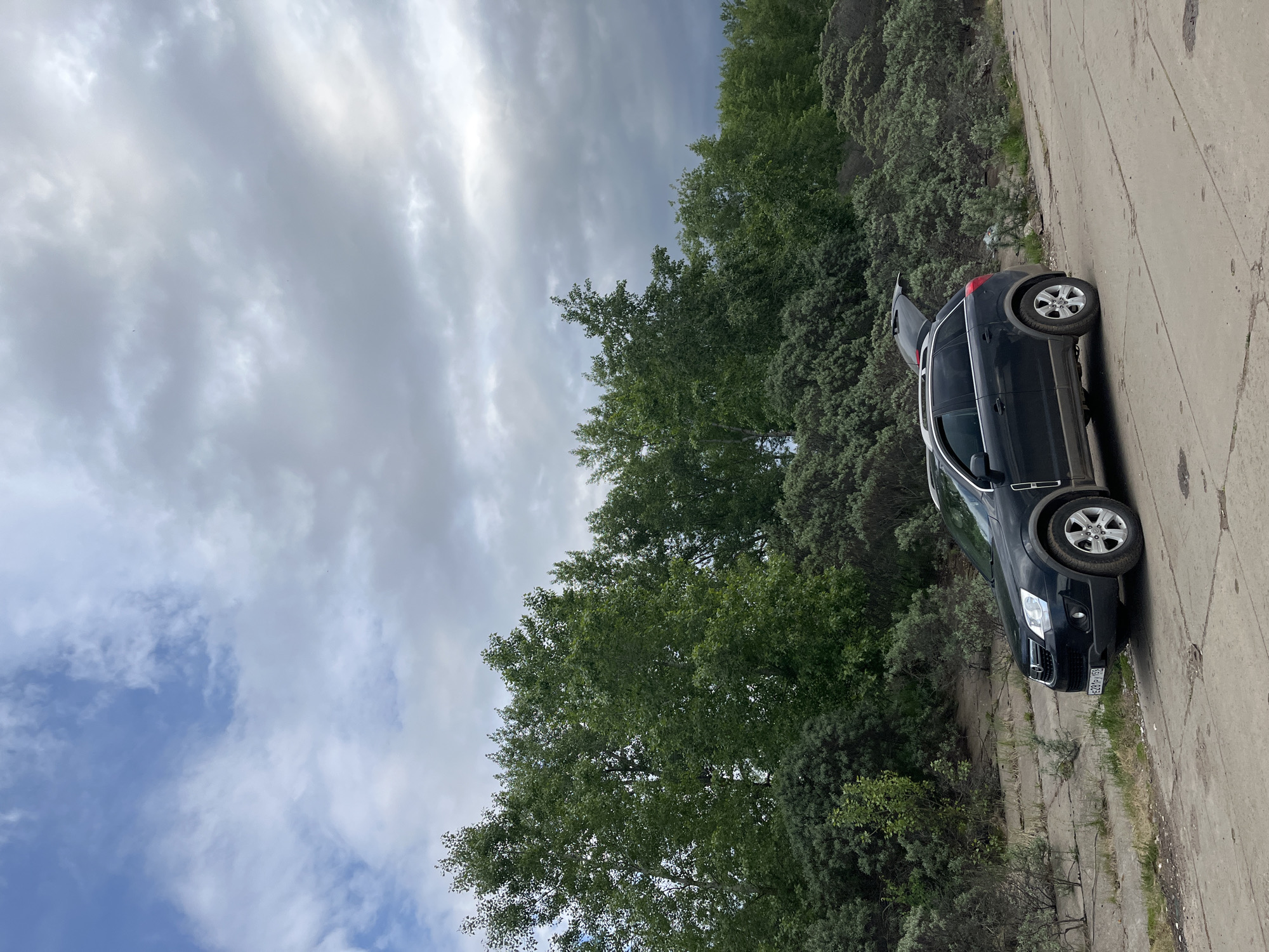 The image presents an outdoor scene with a black SUV parked on a concrete road, likely in a wooded area on the outskirts of a city. The trunk of the SUV is open. The sky is overcast with a mix of gray and white clouds. The vegetation consists of a variety of trees and bushes. There are no people visible in the frame. The vehicle's license plate is partially obscured but appears to contain Russian characters. Based on this information, while a specific location in Chaikovsky, Russia cannot be definitively confirmed without more context, the overall scene is consistent with a location in Russia based on the vehicle's license plate. The environment suggests a rural or suburban setting. The lighting indicates it's daytime, likely under cloudy conditions.