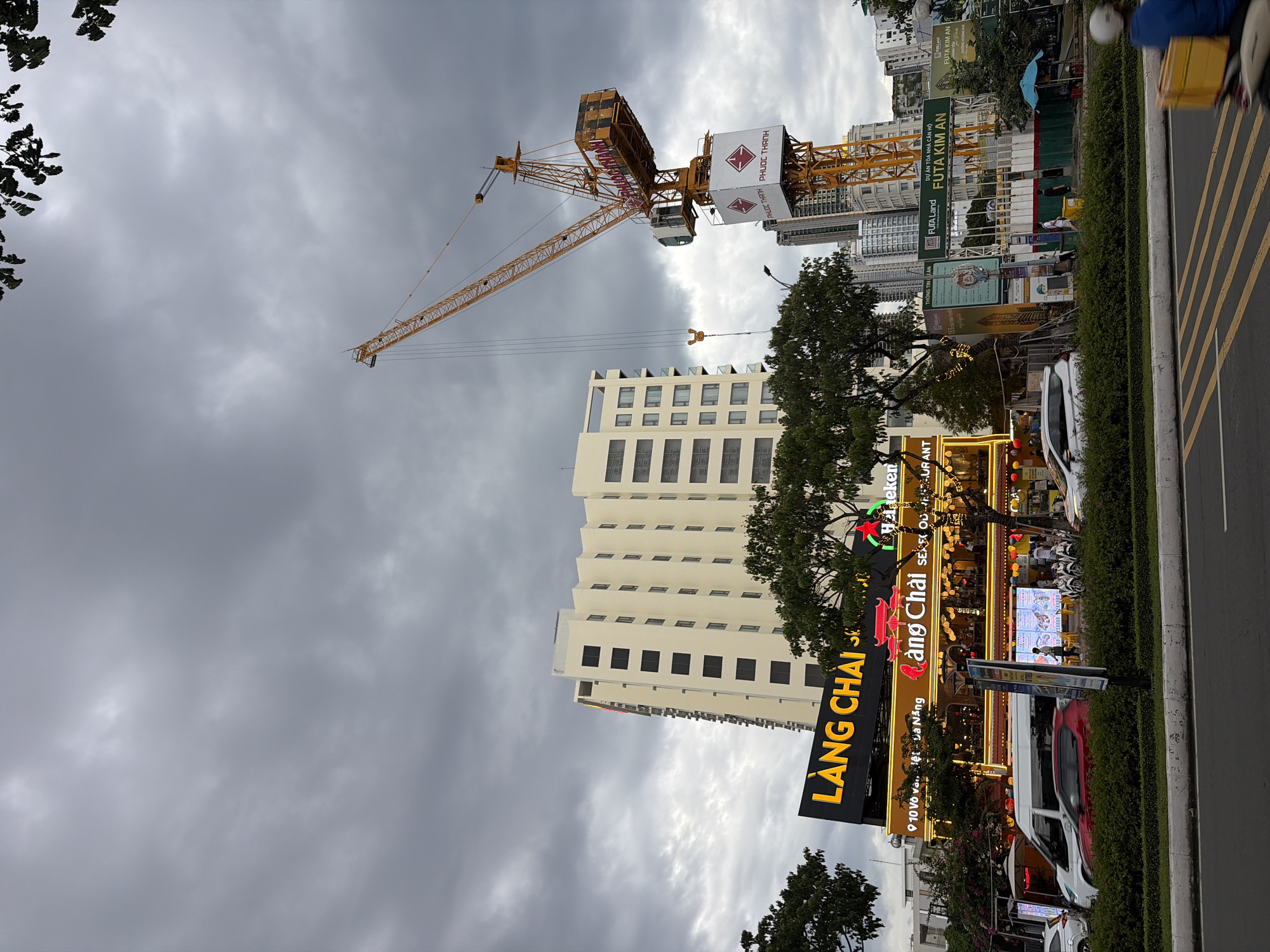 The image shows a street scene in Đà Nẵng, Vietnam, under an overcast sky. A large yellow construction crane dominates the upper right portion of the frame, with its boom extended. To the left of the crane, a tall, cream-colored apartment building with dark rectangular windows stands prominently. In the foreground, a busy street with moving vehicles, including cars and a scooter rider in the lower right, is visible. A row of lush green bushes lines the sidewalk. On the left side of the street, a brightly lit restaurant called "LANG CHAI SEAFOOD RESTAURANT" is identifiable by its illuminated yellow signage and glowing red lanterns. Various other buildings, including some under construction with signage that includes "FUTA Land" and "FUTA KIM AN," are visible in the background, suggesting an active urban development area. The overall atmosphere is that of a city in the midst of construction and commercial activity.