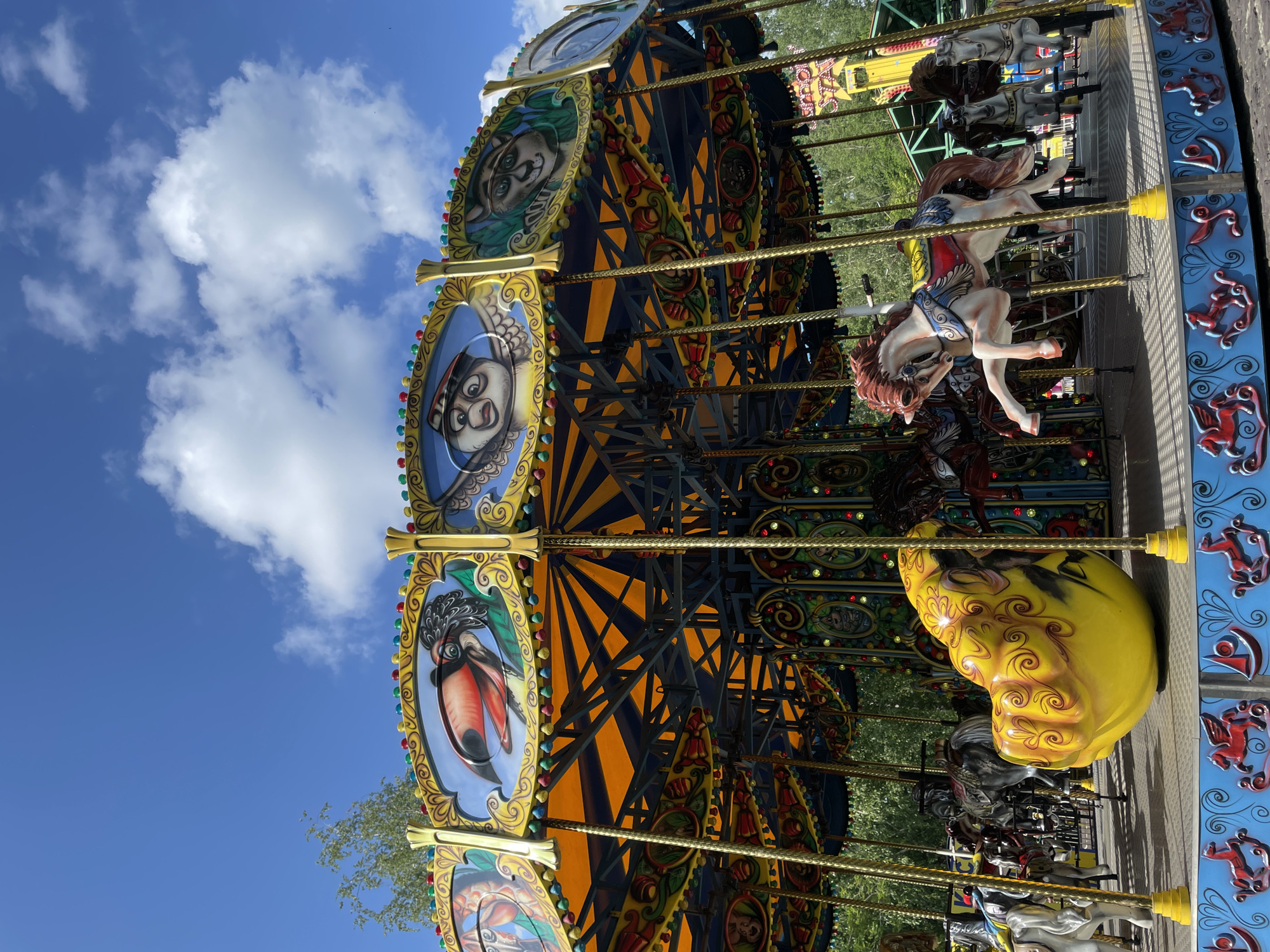 The image shows a carousel at an amusement park on a sunny day. The carousel is adorned with vibrant colors and intricate designs, featuring painted portraits of various characters within oval frames. Among these portraits are images of a character with a red beak and feathers, a playful-looking monkey, and another with a serious expression. The carousel itself is decorated with yellow and blue accents, and ornate gold poles support the structure. Attached to the carousel are several horse-shaped seats, with one prominently displayed in the foreground, a white horse with a reddish-brown mane and tail, detailed with blue and red accents. Other horses of various colors, including gray and white, are visible further back. The background reveals more of the amusement park, with hints of other rides and lush green trees, suggesting a warm, outdoor setting. The sky above is a bright blue with scattered white clouds, indicating pleasant weather. The overall scene evokes a sense of fun and excitement typically associated with amusement parks.