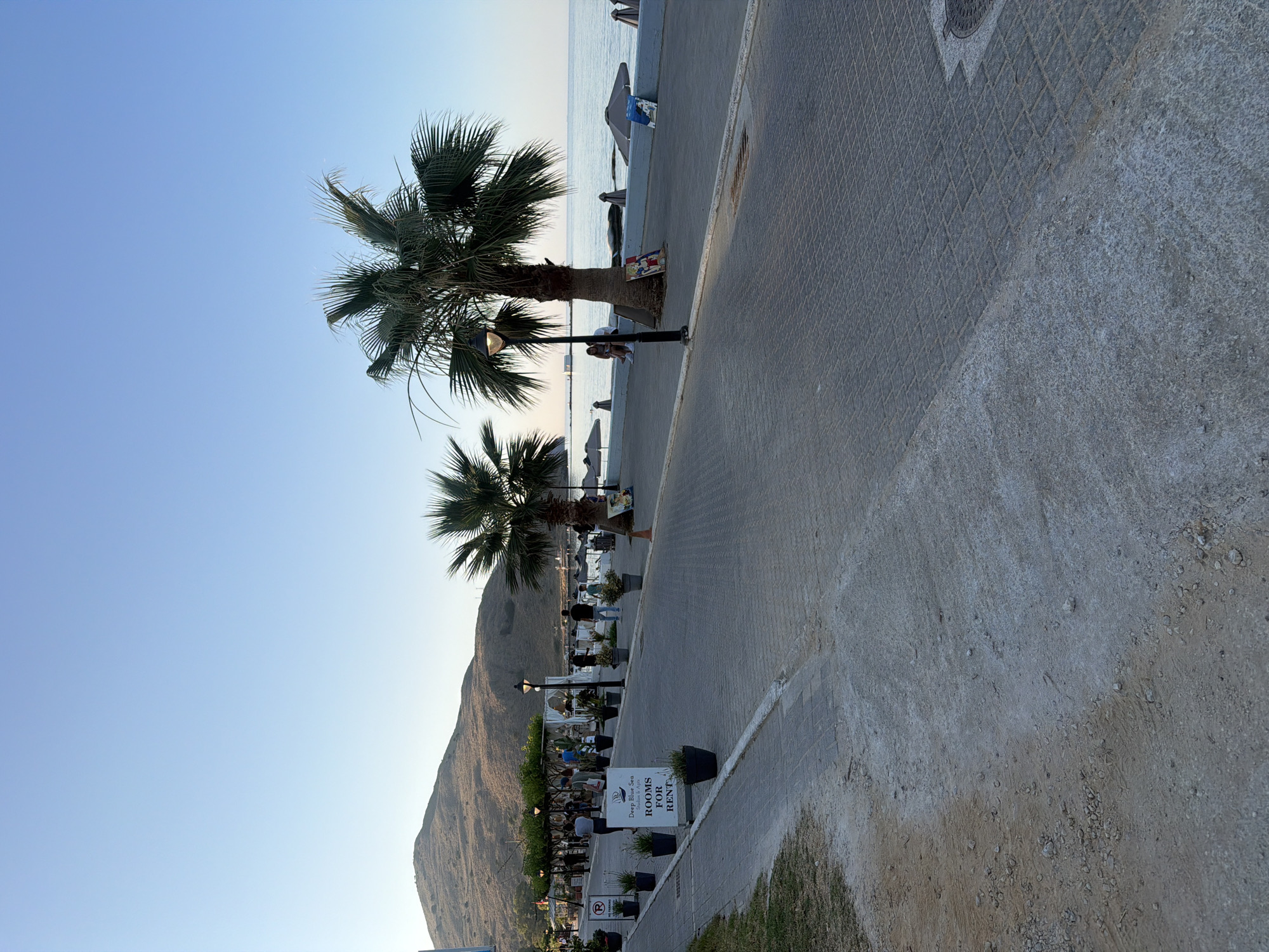 The image shows a pedestrian walkway in Georgioupoli, Greece, on a clear day with a bright blue sky. A paved pathway runs parallel to the coastline, which is visible on the right side of the frame. Several palm trees are planted along the walkway. In the background, a large, barren hill dominates the landscape.
On the walkway, there are several people strolling. Some are seen sitting on benches near the beach. There are also a few signs, one of which reads "Deep Blue Sea Studios & Apartments ROOMS FOR RENT". Beach umbrellas and chairs are set up along the shore. A lamppost stands near a palm tree. The overall scene suggests a typical summer day in a tourist resort town.