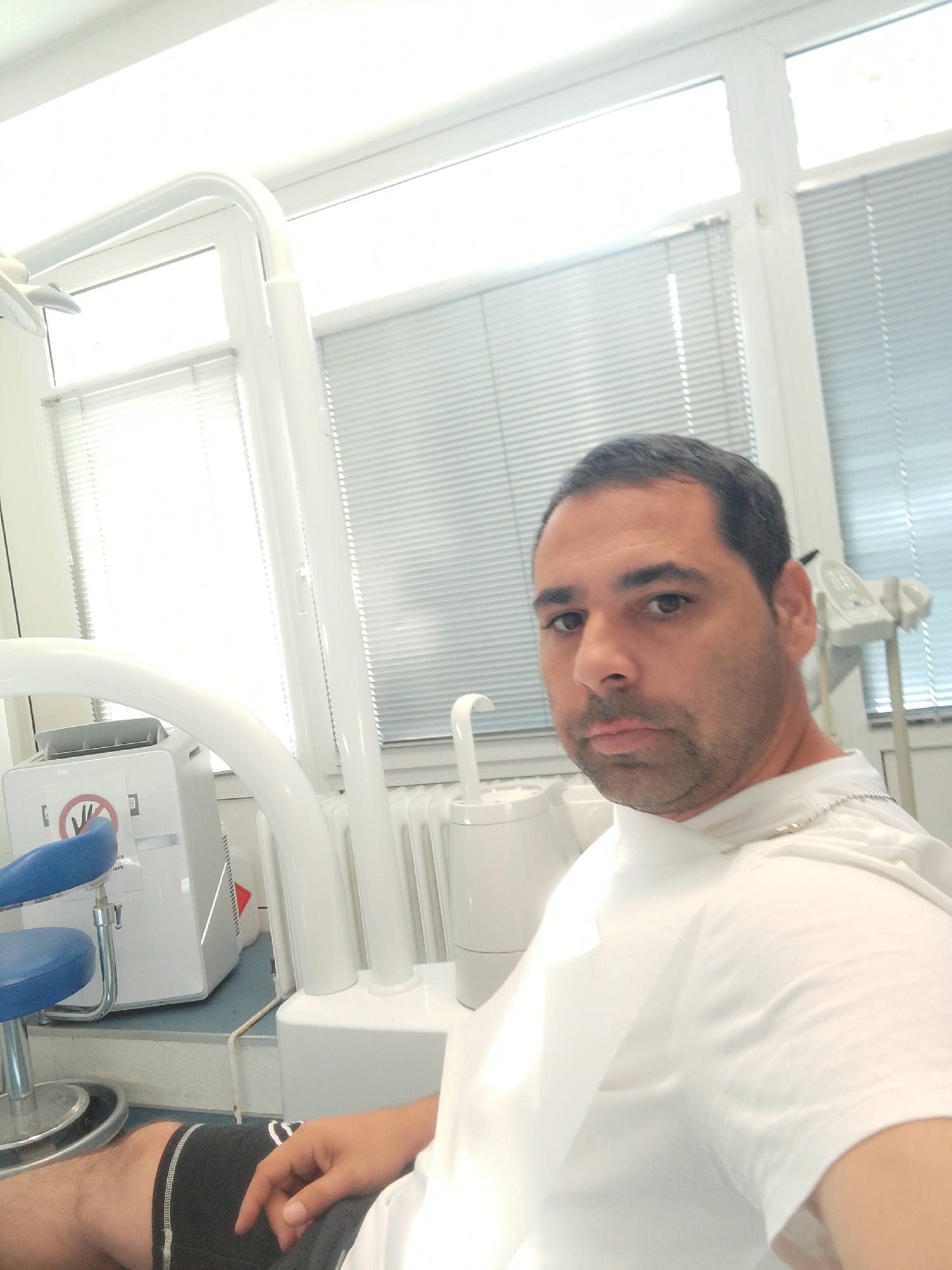 A man is sitting in a dental chair in what appears to be a dental office in Delmenhorst, Germany. He is wearing a white t-shirt and black shorts. The man is looking at the camera and has a neutral expression. The dental chair is blue and chrome. There is a dental unit in the background with various instruments and equipment. The room has large windows with blinds. The overall lighting is bright, suggesting it is daytime.