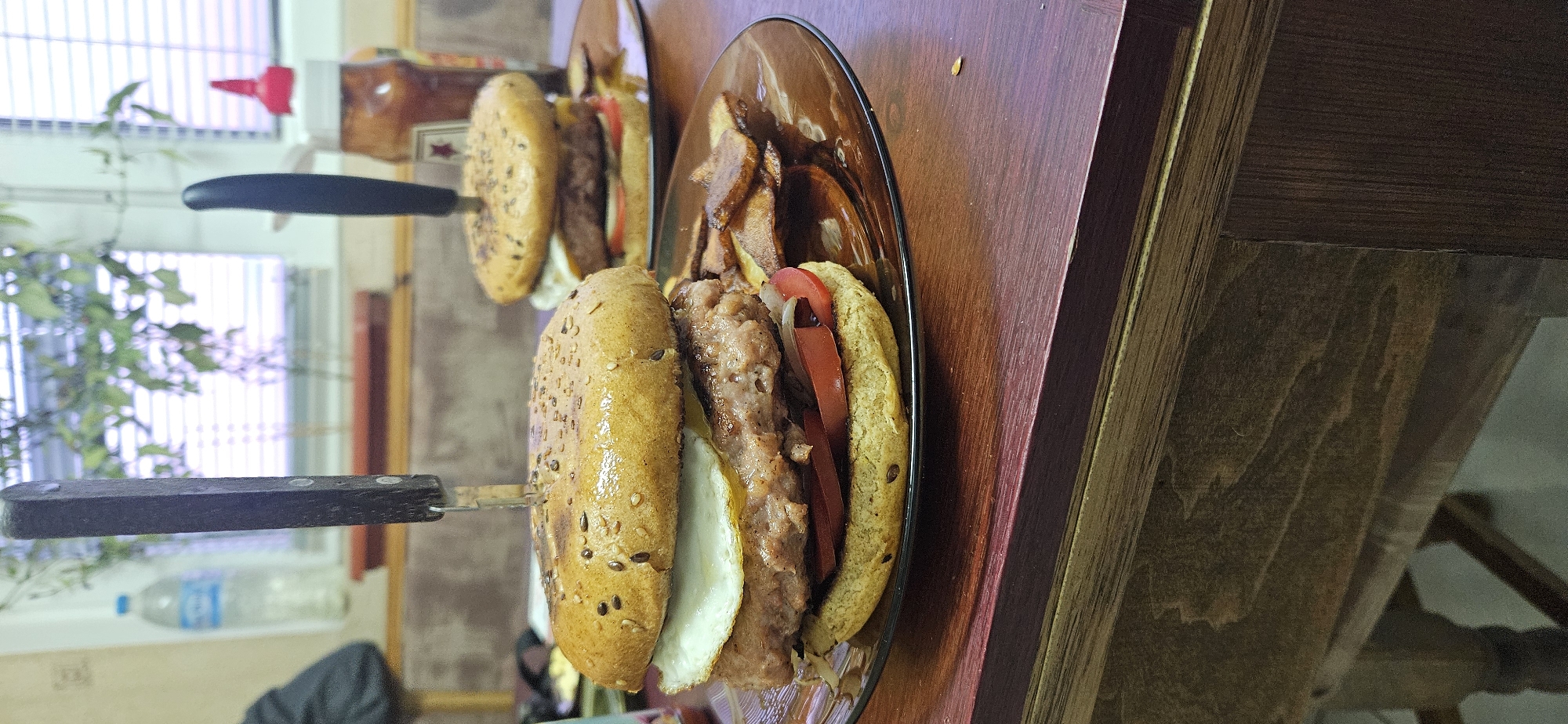 The image shows two plates with sandwiches impaled by knives. The sandwiches appear to have eggs, meat, and tomatoes. French fries are on the plate with the closer sandwich. The sandwiches are on a wooden surface, likely a table. Behind the sandwiches, there is a window with a partially visible plant and a red condiment container. A water bottle is visible in the lower left corner. The location and city are not discernible from the image. The image is oriented sideways.