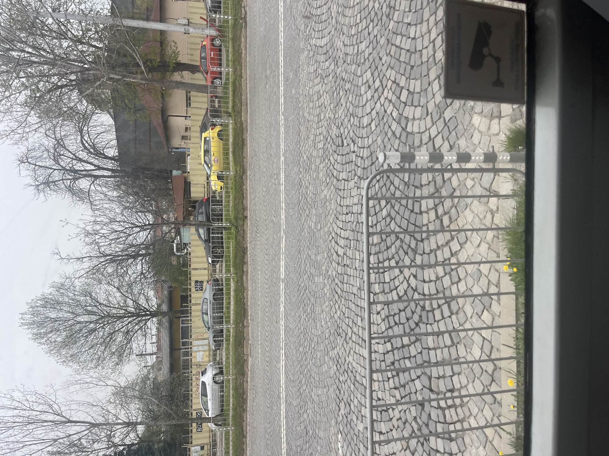 The image depicts a street scene in Sofia, Bulgaria, viewed through a window obscured by a security sign and metal fence. The street is paved with cobblestones and has a double white line dividing traffic. Cars are parked along the left side of the street, including a red car, a yellow taxi, and several silver and black vehicles. Buildings line the street beyond the parked cars, and bare trees are visible. The sky is overcast. The cobblestone road in the city of Sofia, Bulgaria is made of grey stones laid in an arc pattern.