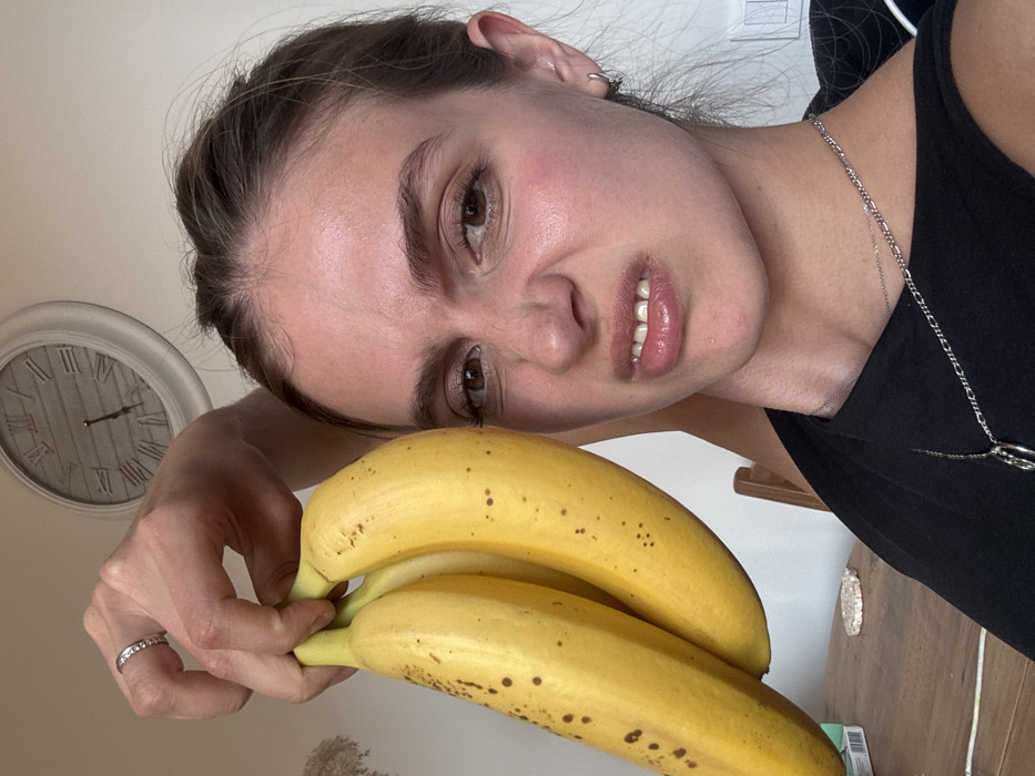 The image displays an indoor scene in an unknown location within the city of Unknown, Unknown. An adult female, positioned in a close-up, selfie-style shot, occupies the majority of the frame. Her brown hair is pulled back, and she exhibits a grimacing facial expression, characterized by furrowed eyebrows, a wrinkled nose, and a partially bared upper lip revealing her teeth. She wears a dark-colored top, two silver necklaces (one a plain chain, the other with a circular pendant), and a small silver hoop earring in her left ear. A silver ring with multiple stones is visible on her left ring finger. Her right hand is raised, holding a bunch of three ripe, yellow bananas with brown speckles, positioned to the left of her head. In the background, a light-colored wall features a circular white wall clock with Roman numerals, indicating approximately 12:45. A wooden table is visible in the lower left background, displaying a white, circular, textured item and a partially visible white and teal box. A wooden chair is also partially visible behind the female.