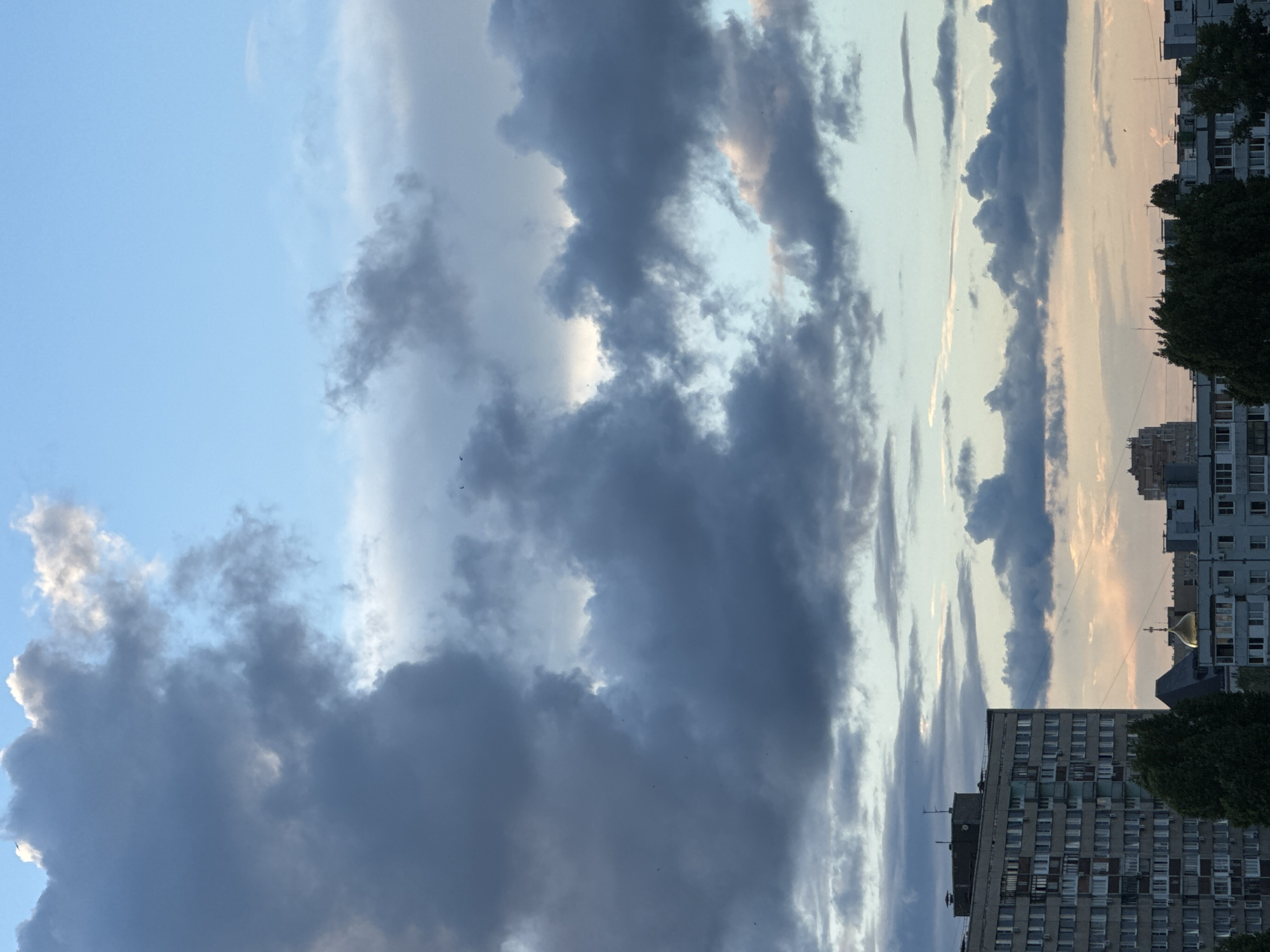 The image captures a cityscape with a dramatic sky. In the foreground, a tall, imposing apartment building dominates the left side, its facade a grid of windows and balconies. To the right of this building, a cluster of other buildings forms the urban silhouette, one of which features a golden dome, suggesting a religious structure. Dark green trees partially obscure the lower portion of the buildings, adding a natural element to the urban scene.
The sky is the main subject, filled with an array of clouds. Large, dark, and heavy clouds occupy the upper portion of the frame, interspersed with lighter, wispy clouds. The lower part of the sky shows a gradient of colors, from pale blue to soft pink and orange hues, indicating either dawn or dusk. The lighting suggests it is either early morning or late evening, with the sun possibly setting or rising behind the clouds. Several small, dark specks in the sky could be birds.