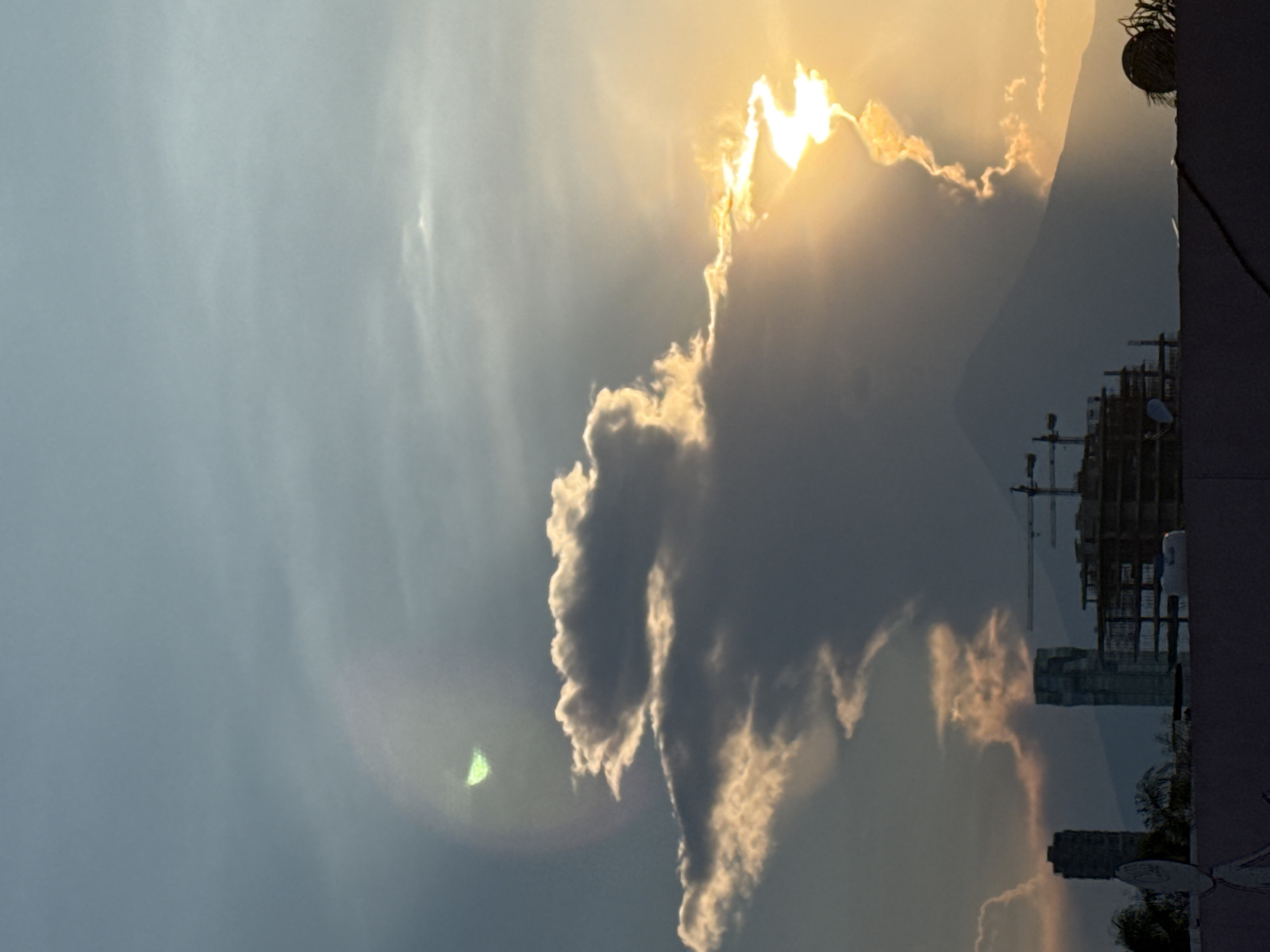The photograph, taken in Puebla, Mexico, depicts a cloud formation partially obscuring the sun. The clouds are dark, with bright, sunlit edges. A large construction site with cranes and a partially completed building is visible in the middle ground. In the foreground, the edge of a building is present, with a hanging planter. In the background is another mountain, also partially obscured by clouds.