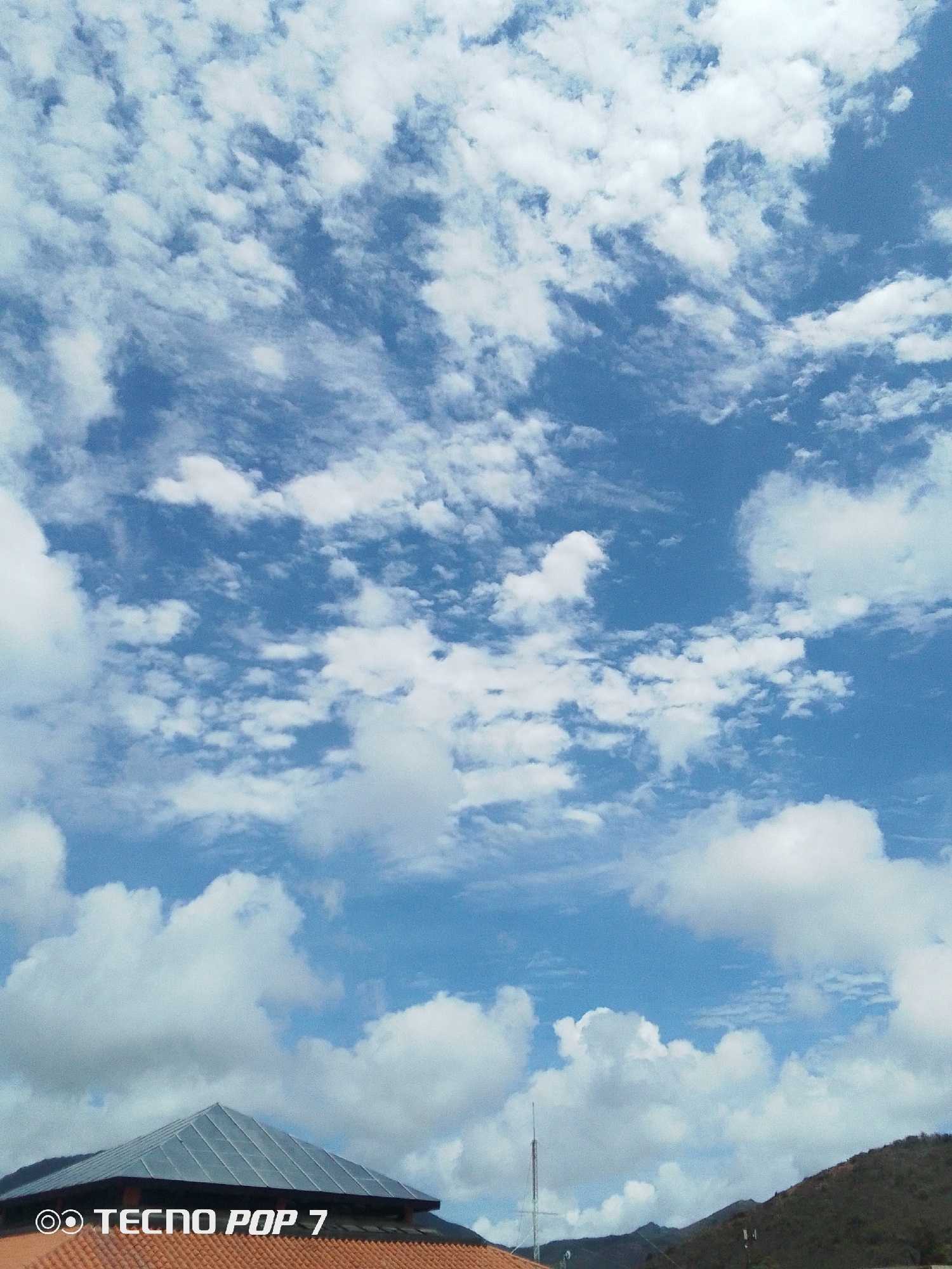 The image shows a view of the sky above Pedro González, Venezuela. The sky is blue with scattered white clouds. In the lower portion of the image, a building with a gray, triangular roof and a red tile roof underneath is visible. Behind the building, a grassy hill slopes upward, and a radio tower is visible. Mountains are also present in the background. The phone used to take the picture is a TECNO POP 7.