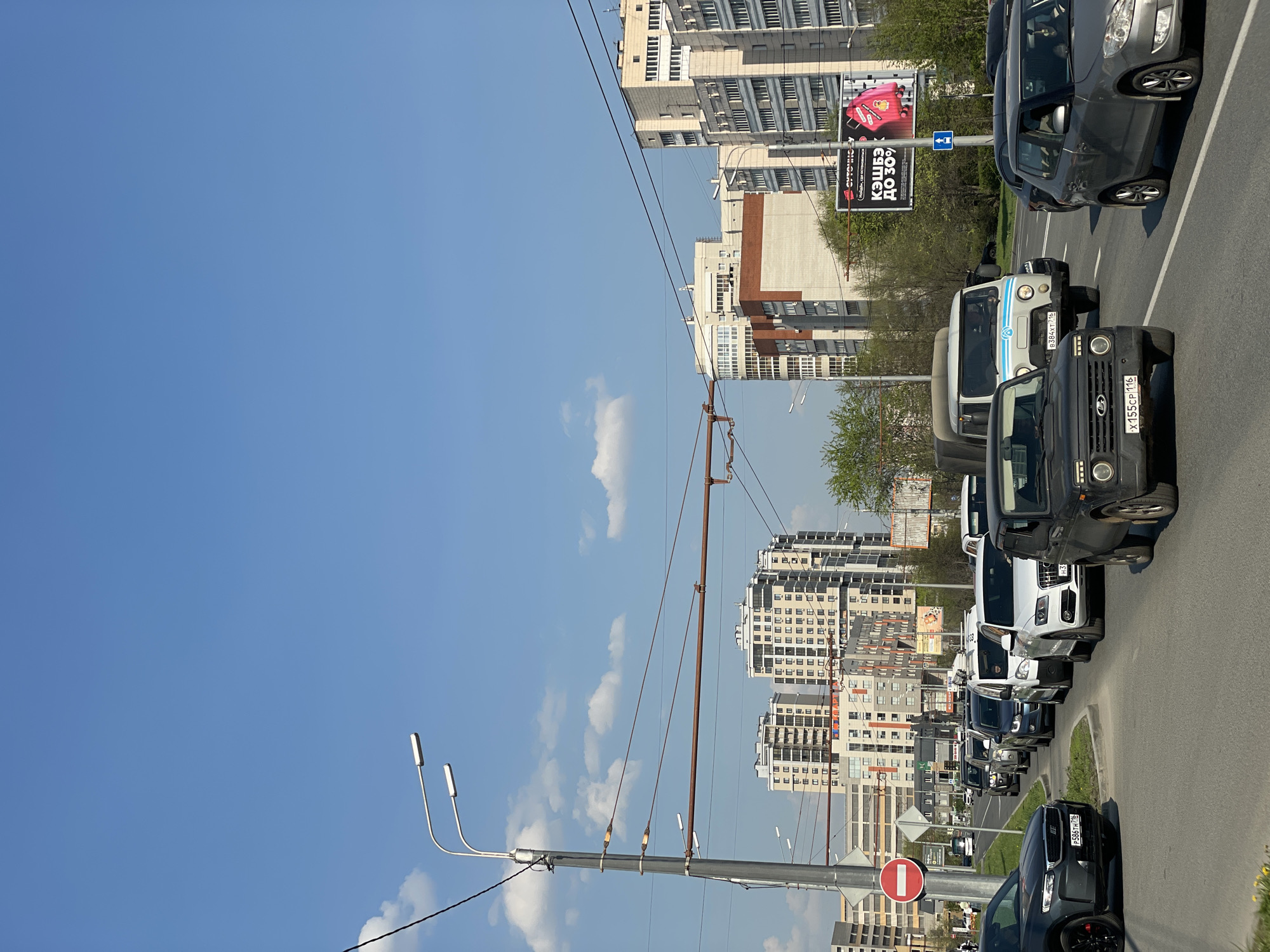The image shows a street scene in an urban area, with buildings visible in the background and a row of cars lined up along the right side of the road. There are power lines and poles visible in the foreground. A billboard advertises "cashback up to 30%". A red and white "Do Not Enter" sign is also visible. The sky is blue with some clouds. The location is in the city of Unknown, Unknown.