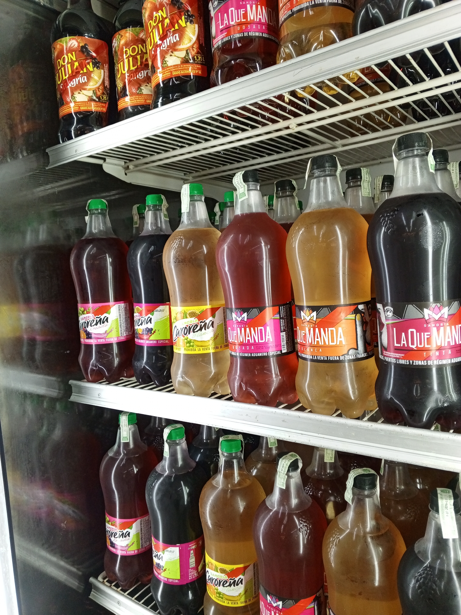 The image shows a refrigerated display of bottled beverages, likely in a store located in Porlamar, Venezuela, based on the labeling of the products. The top shelf features "Don Julian Sangria" bottles. The lower shelves contain rows of "Caroreña" and "La Que Manda" sangria bottles in various flavors (Rosada, Blanca, Tinta), distinguished by different colored labels and liquid hues. The bottles have seals on their caps.