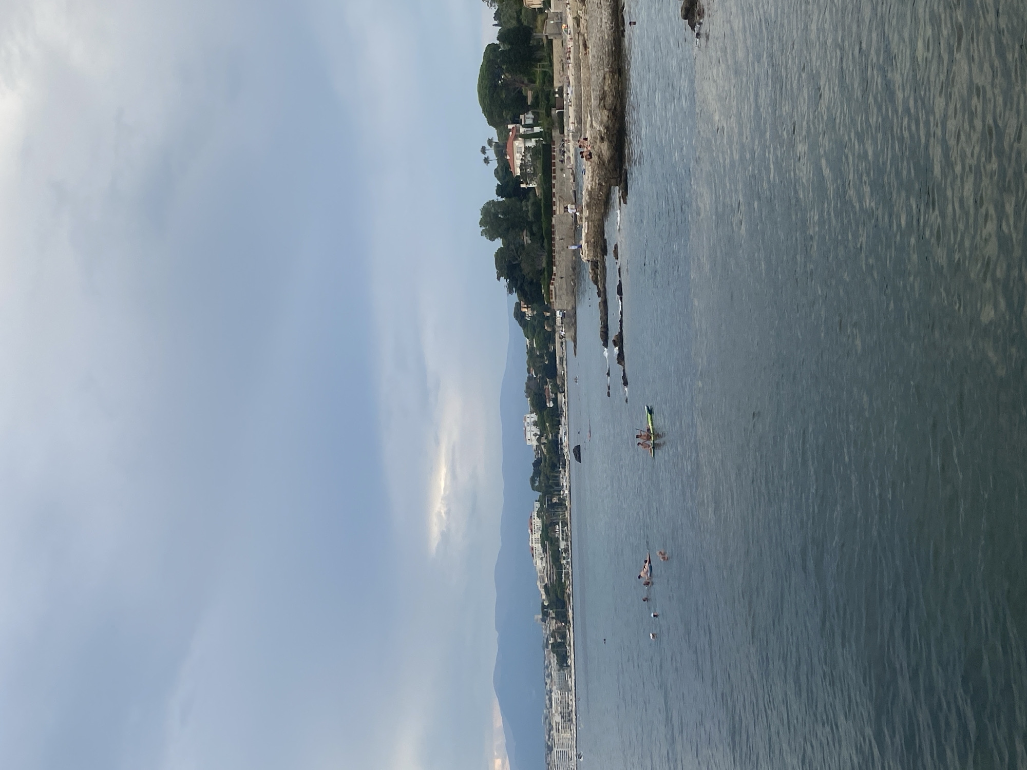 The image depicts a cloudy day at a coastal area in Antibes, France. The foreground is dominated by the deep blue, slightly rippled water of the sea. In the mid-ground, a rocky shoreline features several people sunbathing and walking. A low stone wall runs along the top of the shoreline, with lush green trees and a large white house with a red roof situated behind it. Further in the distance, the coastline is lined with buildings, suggesting an urban area, and receding mountains are visible under the overcast sky. In the water, several individuals are swimming or relaxing, and two people are paddling on a green stand-up paddleboard. The overall scene conveys a sense of leisure and a coastal lifestyle.