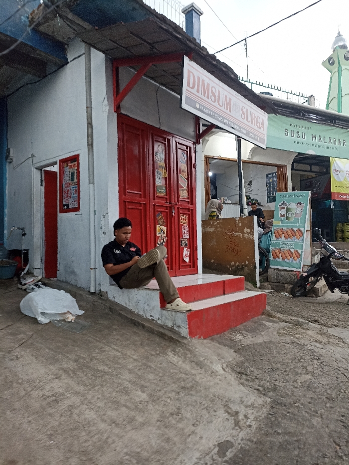 The image depicts a street scene in Jatinangor, Indonesia (latitude: -6.935, longitude: 107.773). A young man in a black shirt and khaki pants sits on the steps of a red-door storefront with a white exterior. The storefront is labeled "Dimsum Surga," a name written in both English and Indonesian. The man is looking down at a mobile device in his hand. There are other storefronts visible, one with a sign advertising "Susu Malabar." An older man wearing a white hat is visible in the background, standing near the storefront with the "Susu Malabar" sign. Another individual can be seen further down the street, standing in front of another shop with the "Susu Malabar" sign. A motorcycle is parked in the street near the "Susu Malabar" storefront. There are two white plastic bags on the ground in front of the young man. There is a partially obscured, white, metal garbage can to the left of the young man. The image has a slightly overcast, hazy quality, indicating a partly cloudy or possibly rainy day. The street surface is comprised of rough grey concrete.