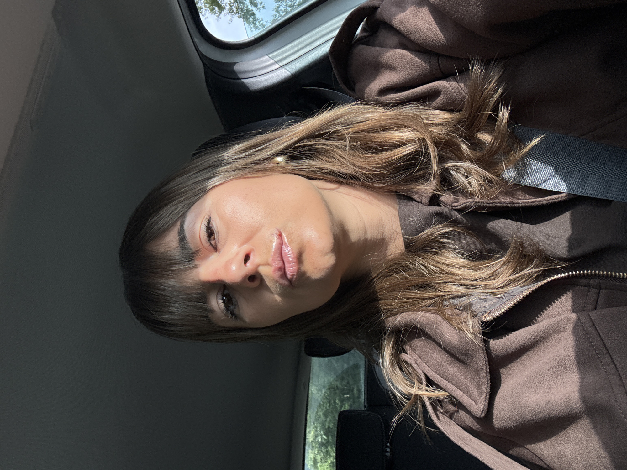 A young woman is sitting in a car. She has brown hair with blonde highlights and brown eyes, wearing a brown jacket and a seatbelt. The car window shows green vegetation and sunlight, suggesting an outdoor location. The scene appears to be taken during daylight. This cannot be identified as being in Mandelieu-la-Napoule, France from the given image and details.