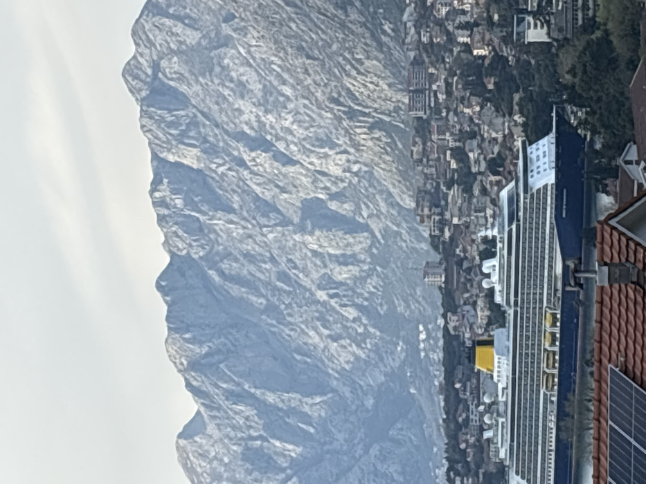 The image displays an elevated, panoramic view of Škaljari, Montenegro, situated along a body of water with a prominent mountainous backdrop. In the immediate foreground, a reddish-brown tiled roof is visible, equipped with multiple dark, rectangular solar panels and a light-colored chimney structure. A large, multi-deck cruise ship, identified by partially visible text on its dark blue hull as "SPIRIT OF DISCOVERY," occupies the mid-ground on calm water. The vessel features a white superstructure with numerous decks, visible windows, a distinctive yellow funnel at its stern, and multiple white radar domes or communication antennae on its upper decks. Behind the ship, the urban area of Škaljari extends from the water's edge up the lower slopes of the mountains, characterized by densely packed residential buildings with red-tiled roofs, interspersed green foliage, and several taller, modern structures. The background is dominated by an extensive, rugged mountain range, exhibiting steep, grey, rocky slopes and multiple peaks, under an overcast, light grey sky. No human figures are discernible within the scene.