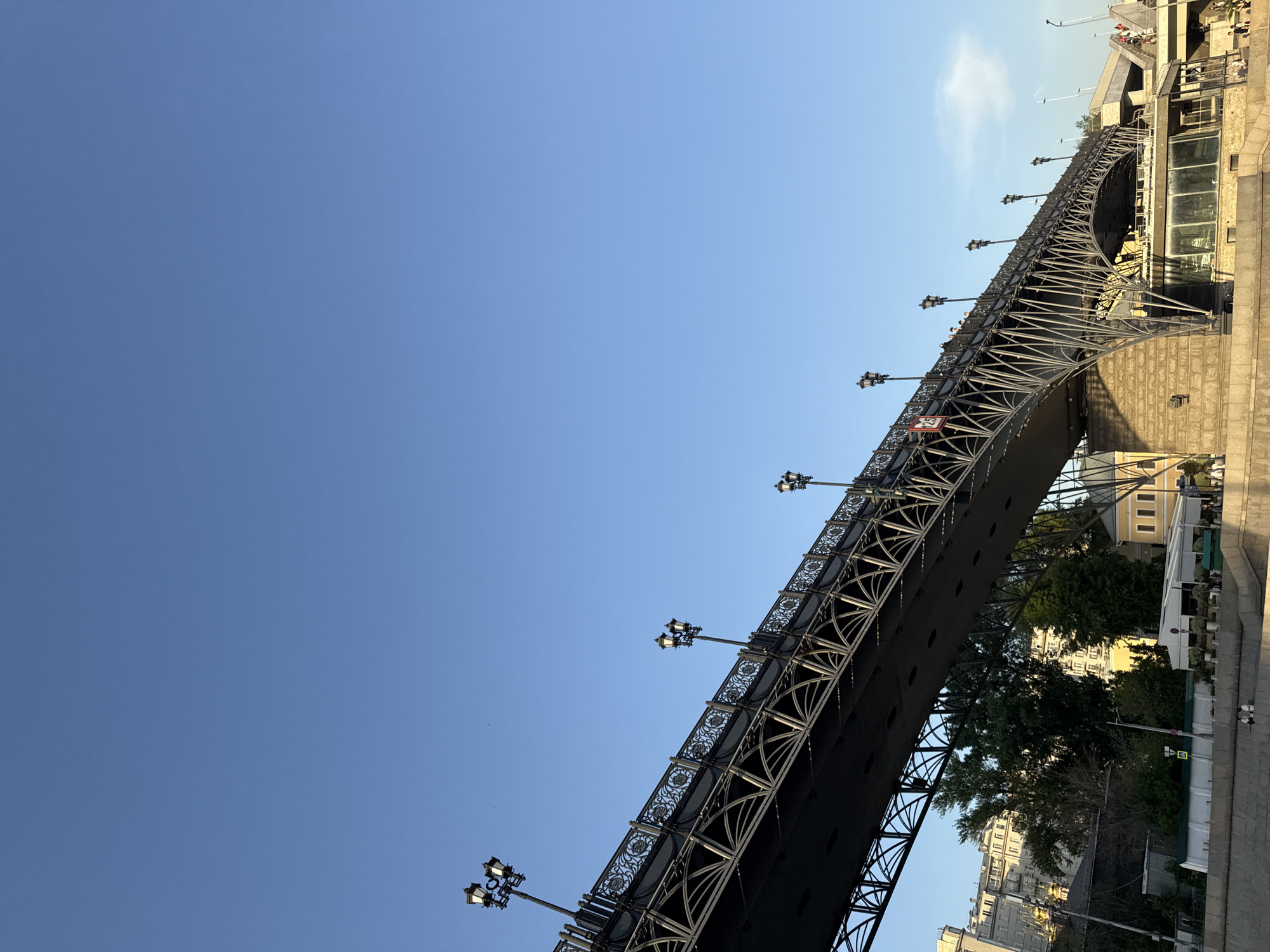 The image shows a view from below of the Bolshoy Moskvoretsky Bridge in Moscow, Russia. The bridge is an arch bridge constructed of steel and has ornamental lamp posts along its length. The sky is clear and blue. Beneath the bridge, trees and buildings are visible along the Moskva River embankment. The bridge carries pedestrian and vehicular traffic. The photograph is taken at an angle, tilting upward.