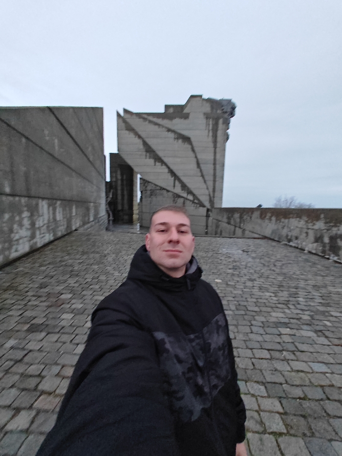 A male subject is captured in a selfie-style image at the Monument to 1300 Years of Bulgaria (Founders of the Bulgarian State Monument) in Shumen, Bulgaria. The subject, a light-skinned male with short, buzz-cut hair, wears a black hooded jacket with a dark grey camouflage pattern across the chest and upper sleeves, and a grey inner collar. He faces the camera directly, holding the device at arm's length, and exhibits a faint smile.
Behind the subject, the distinctive Brutalist architecture of the monument dominates the background under an overcast sky. Large, angular concrete slabs form imposing structures. To the left, a tall, flat concrete wall-like section is visible, featuring subtle diagonal lines. To the right, another large concrete structure rises, characterized by sharp, geometric facets and visible water stains or weathering streaks running vertically. A dark, narrow opening or passageway is discernible between these two main sections. The ground is paved with irregularly shaped grey cobblestones. In the far distance on the right, sparse, bare tree branches are visible against the muted sky, indicating a possibly cool or cold outdoor environment.