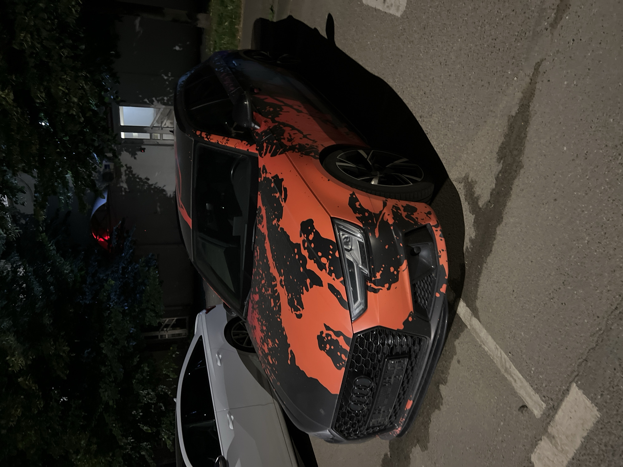 A custom-wrapped Audi RS4 Avant is parked in a lot at night. The car has a black and orange splatter-paint design. A white Audi A6 is parked behind it. The background shows trees and a building with an open doorway. The scene appears to be a parking lot in Krasnodar, Russia.