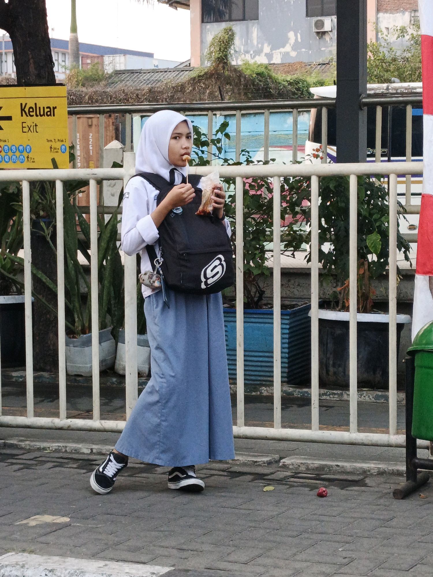 A young Muslim woman wearing a white hijab, a white school uniform shirt, and a long blue skirt walks along a sidewalk next to a white metal fence. She has a black backpack with a white "S" logo on it slung over her left shoulder and is eating a snack from a small plastic bag. She is wearing black Vans sneakers. To the left of the woman, a yellow sign with black text reads "Keluar" and "Exit." The background shows buildings and greenery, including a large tree on the far left. The scene appears to be in a public area, possibly a school or transport hub, given the presence of the "Exit" sign and fence. The ground is paved with gray interlocking bricks. A green trash can is visible on the right side of the frame. The woman's facial expression is neutral as she looks towards the camera.