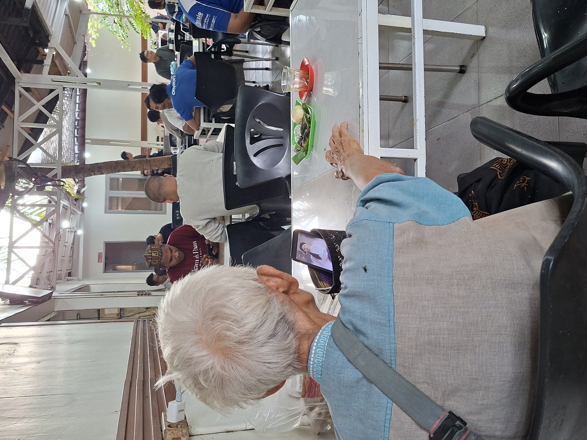 The image depicts an indoor scene in Lhokseumawe, Indonesia, where several individuals are gathered, possibly in a cafe or public eating area. An elderly man with white hair is in the foreground, viewed from behind, using a mobile phone. A table with food and drinks, including a traditional Indonesian snack in a green container and a glass of drink, is visible in front of him. Other people are seated at tables in the background, some of whom are eating and conversing. The building structure shows a mix of traditional and modern elements.