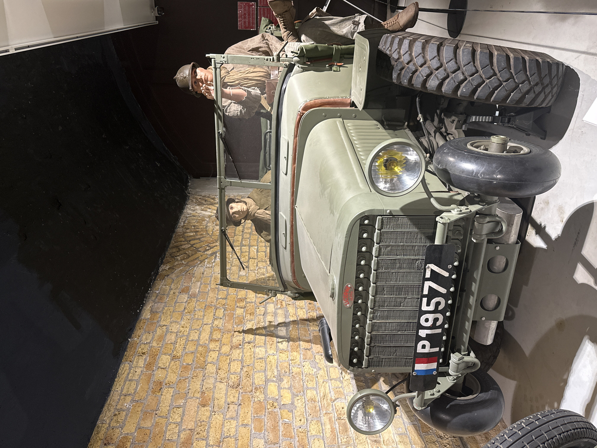 An outdoor exhibit in Dunkirk, France features a vintage military jeep with two mannequin figures dressed in military attire. The jeep is olive green with a prominent grille and two large headlights. A license plate reading "P19577" with French flag colors is affixed to the front. The jeep is parked in front of a textured brick wall with a dark, arched ceiling overhead. The mannequins are positioned within the jeep; one is driving, and the other appears to be observing something ahead. Both mannequins wear helmets and scarves, evoking a wartime setting. The overall presentation suggests a historical display related to the events of Dunkirk.