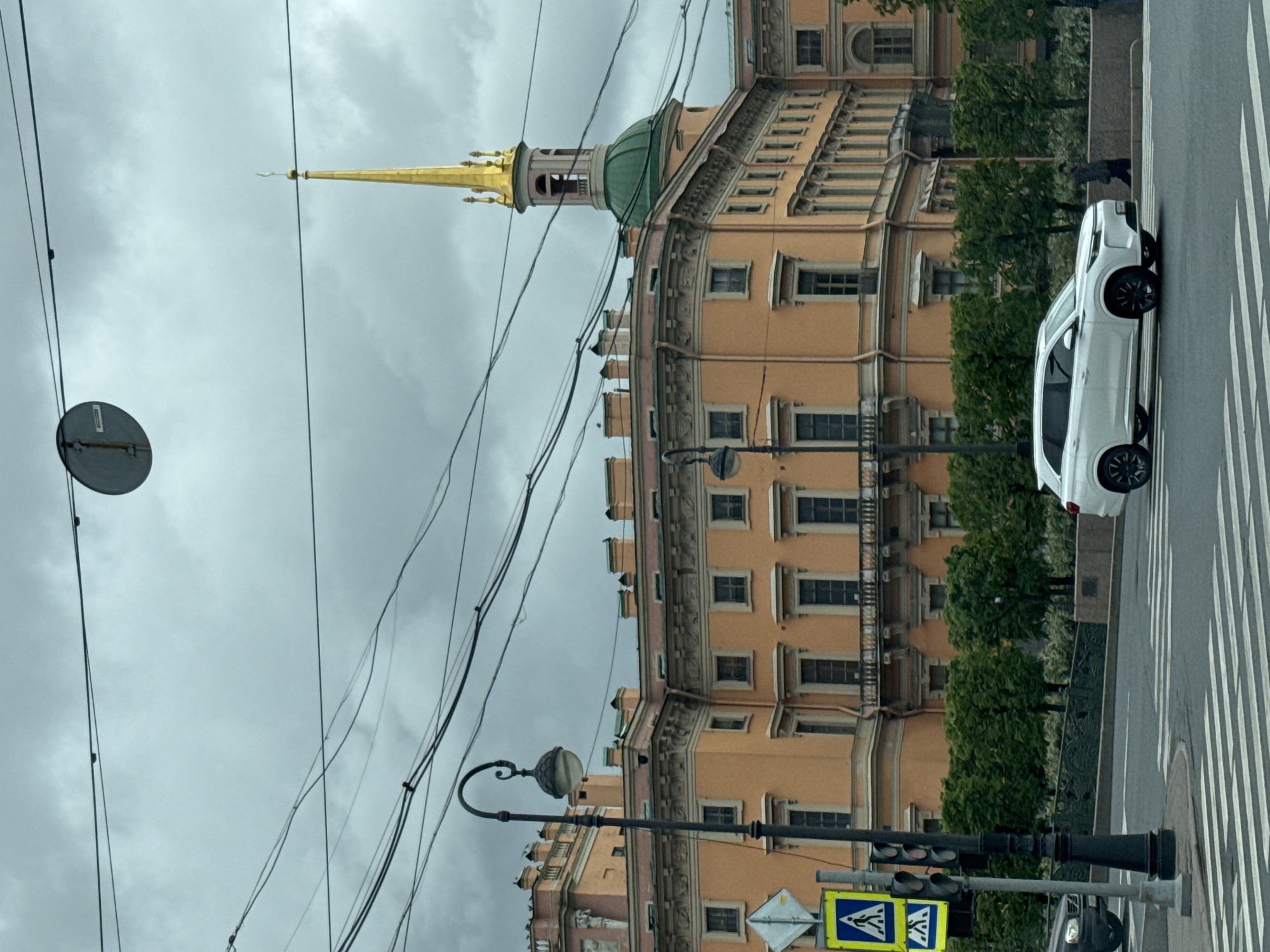 The image depicts a street scene in Saint Petersburg, Russia, dominated by a historical building with a golden spire and a green dome. The building is peach-colored, with symmetrical rows of windows and ornate architectural details. An overhead view shows an assortment of utility wires crisscrossing the sky above the building.
To the right of the building is a crosswalk with white stripes where a white compact SUV is parked along the side of the street. Further along the road, a pedestrian is walking.
In the foreground, there is a street lamp post with a spherical lamp and a traffic light. A pedestrian crossing sign is attached to the lamppost. The sky is overcast and grey, suggesting a cloudy day.