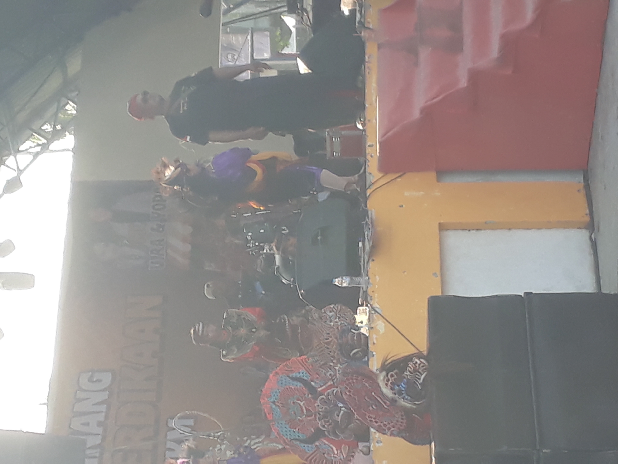 The image depicts an outdoor stage performance in Pangkalpinang, Indonesia. In the background, a banner with Indonesian text, partially obscured and faded, is visible. A few performers are on stage, some dressed in traditional attire. One individual in the foreground is wearing a striking red and blue feathered costume, possibly a headdress or part of a puppet. Another performer in a purple and yellow outfit stands to the right, while a man in a black shirt and red head covering is also present. The stage itself has a yellow base with red stairs on the right. Speakers and audio equipment are visible, suggesting a musical or theatrical event. The overall lighting suggests it is daytime, with sunlight filtering through an overhead structure. The environment appears to be an open-air venue, possibly a public square or event space within the city.