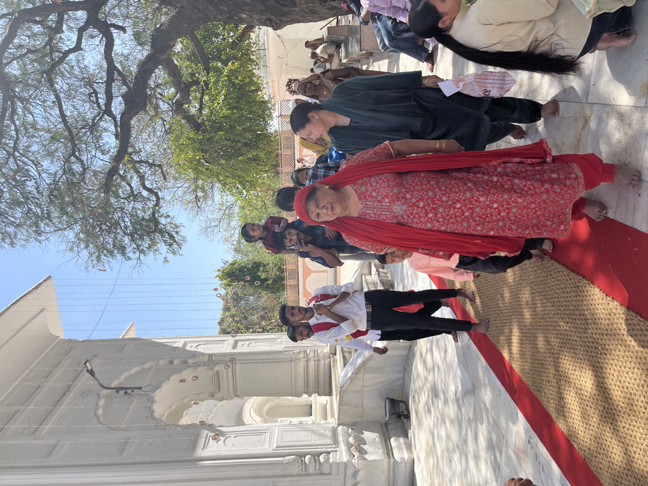 The image shows a group of people walking on a red and beige carpet leading towards a white building. The building appears to be a religious temple or a place of worship, with ornate decorative elements on the walls and a large archway. The people are dressed in traditional Indian attire, with the woman in the center wearing a bright red salwar kameez and a matching dupatta. A younger child is being carried on the shoulders of a man, who is walking behind the woman. The man in the center is wearing a white shirt and black pants, with a black belt. He is holding his hands in a prayer-like position. Other individuals are walking on either side of the woman. There is a large tree in the background, providing shade. The scene is likely set in the grounds of a religious complex. The coordinates suggest the location is in Sector 12, Panchkula, Haryana, India. The individuals are likely participating in a religious procession or ceremony.