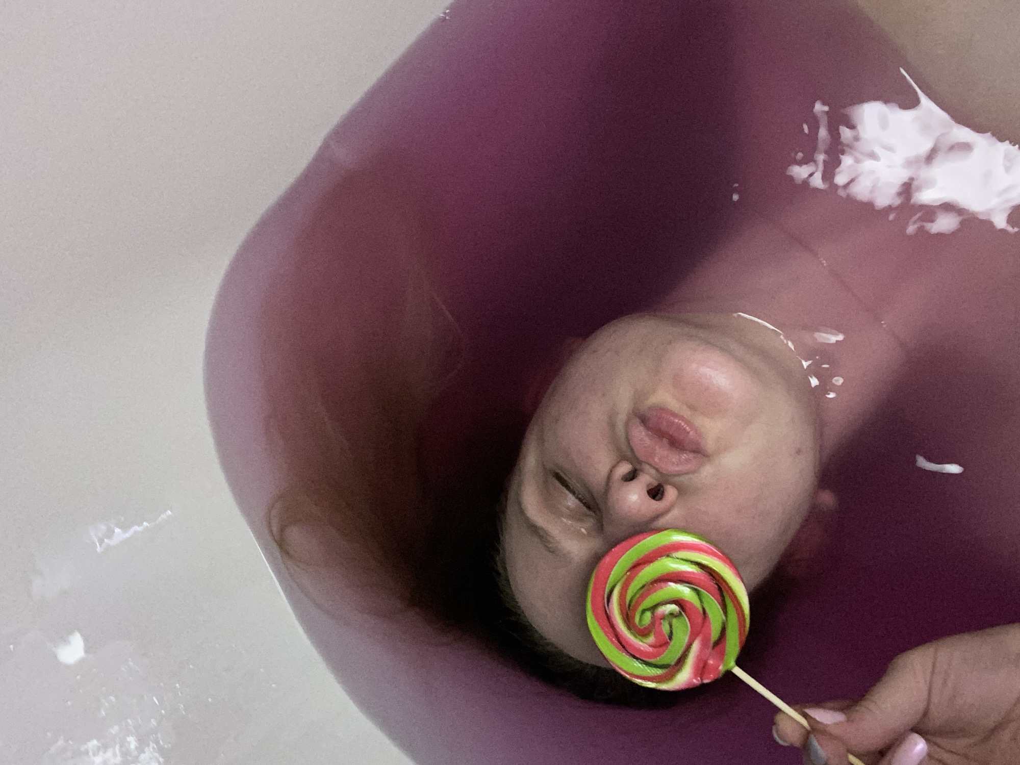 A person is lying in a purple-hued bath with a lollipop held over their closed eye. Their lips are pursed. The hand holding the lollipop is visible in the lower left corner of the image. The person's hair is spread out in the water. The scene is indoors, within a bathtub. The location is unknown.