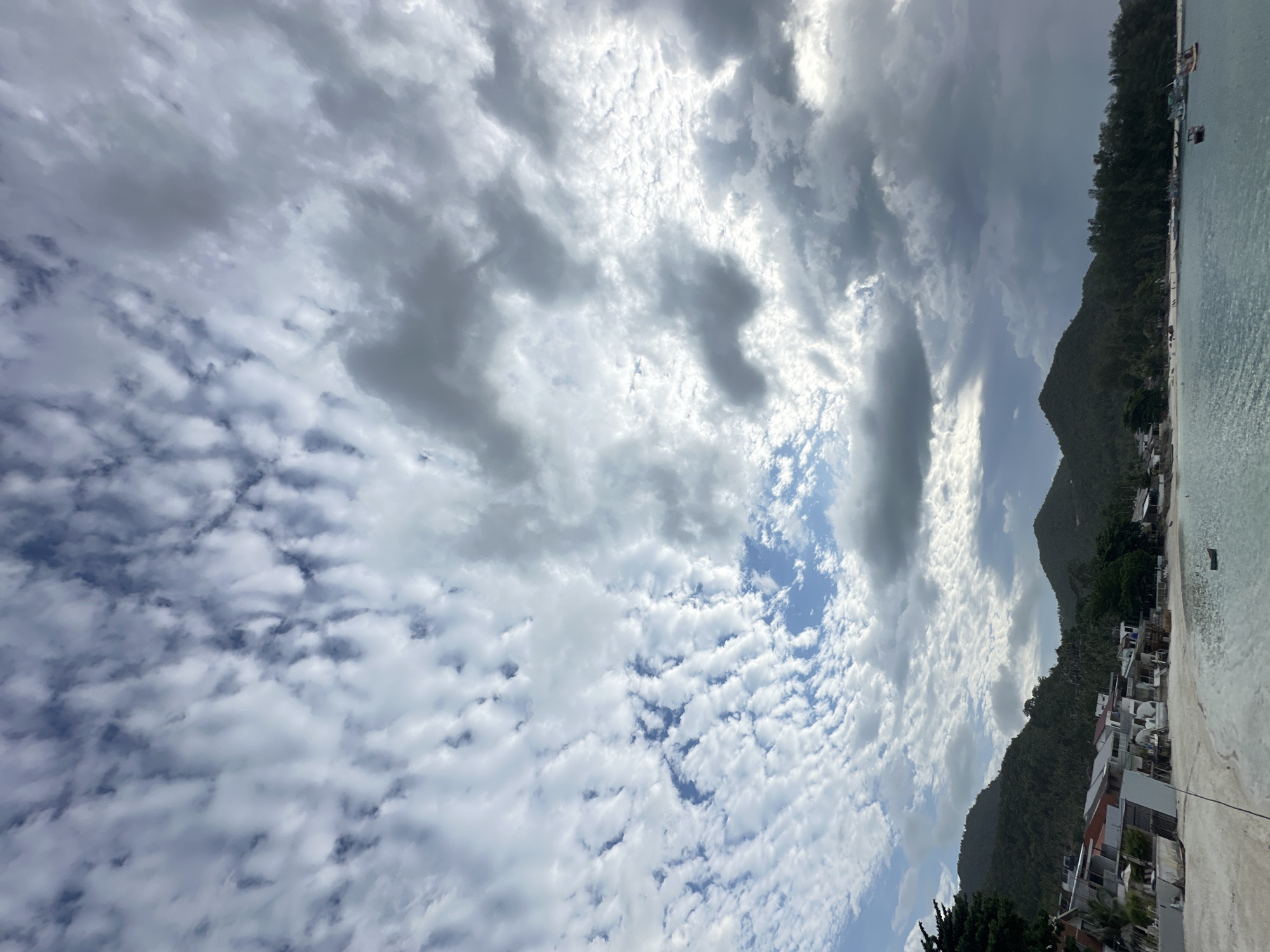 The image depicts a scenic view of a beach and coastline in Tambon Ko Phangan, Thailand, under a dramatic sky. The foreground features a tranquil, light-blue sea with a few small boats visible. A white sandy beach lines the coast, with a scattering of buildings, possibly resorts or homes, situated along its edge. Behind the beach, lush green hills rise, covered in dense vegetation. The sky above is filled with a complex cloud formation, with patches of bright blue sky peeking through, contrasting with darker, heavier clouds, suggesting a dynamic weather system. The overall impression is one of natural beauty and a coastal settlement.