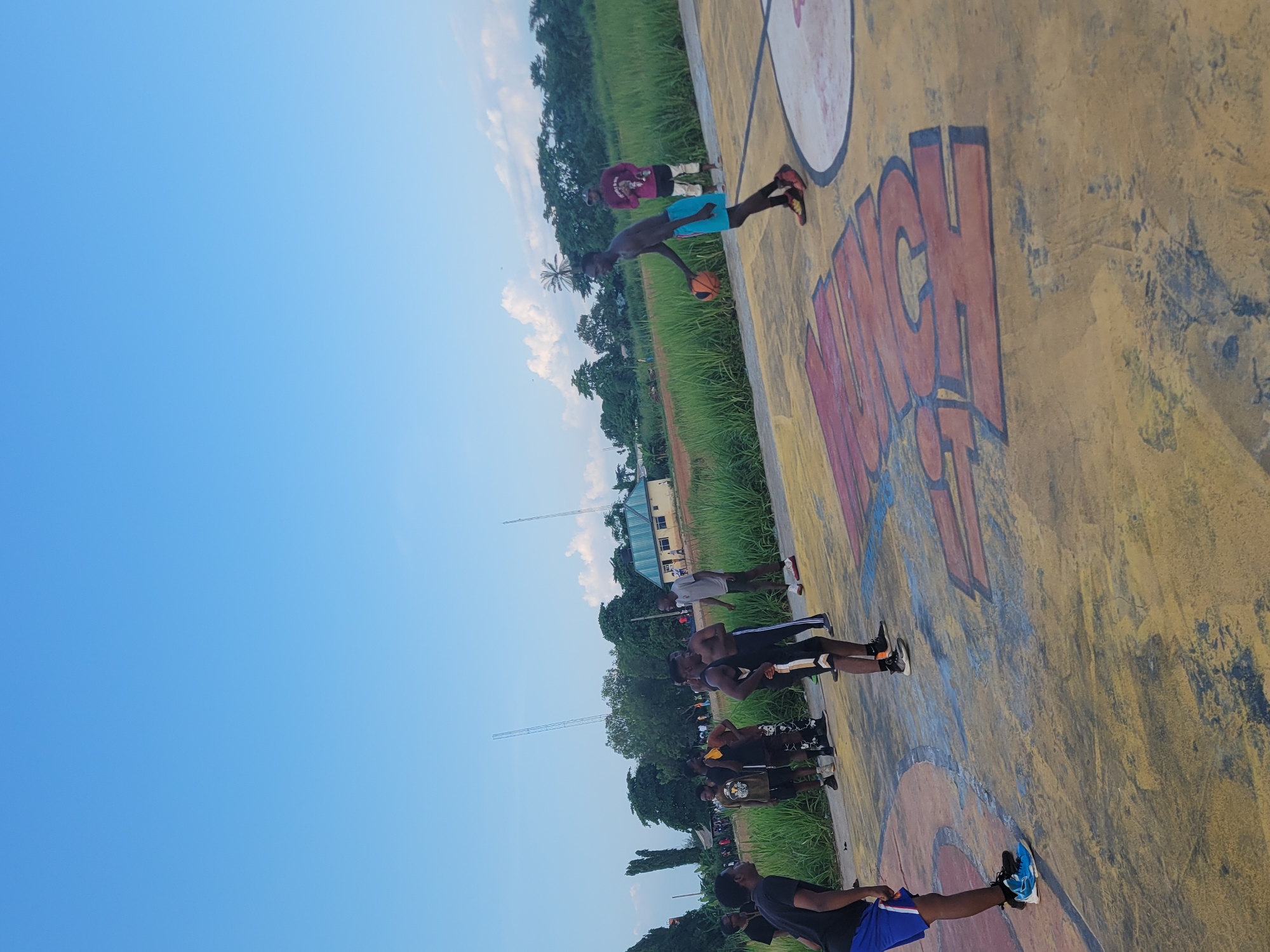 This media file captures an outdoor basketball court scene in Egbagu, Nigeria, under a bright, clear blue sky with scattered clouds, suggesting daytime. The court itself is worn and textured, painted with vibrant yellow and accented with large, red, graffiti-style lettering that appears to spell out "LUNCH." The scene is populated by a group of young people engaged in basketball activities.
In the foreground, a person in blue shorts and a black t-shirt is holding a basketball, seemingly in the process of playing or about to play. To their left, a line of individuals, most of them young men, are positioned in a row, looking up as if participating in a drill or waiting their turn. They are dressed in casual athletic wear, including t-shirts, shorts, and sneakers. Further back, two individuals are visible near the edge of the court, one in a maroon shirt and the other in a dark shirt and shorts.
The background is characterized by lush green grass and trees, hinting at a natural, possibly rural or semi-rural, environment surrounding the court. Some distant buildings and what appear to be communication towers are also visible. The overall impression is one of casual recreation and community engagement, with the bright lighting and clear sky suggesting pleasant weather for outdoor activities. The energy of the scene is palpable, with the focus on the game and the participants.