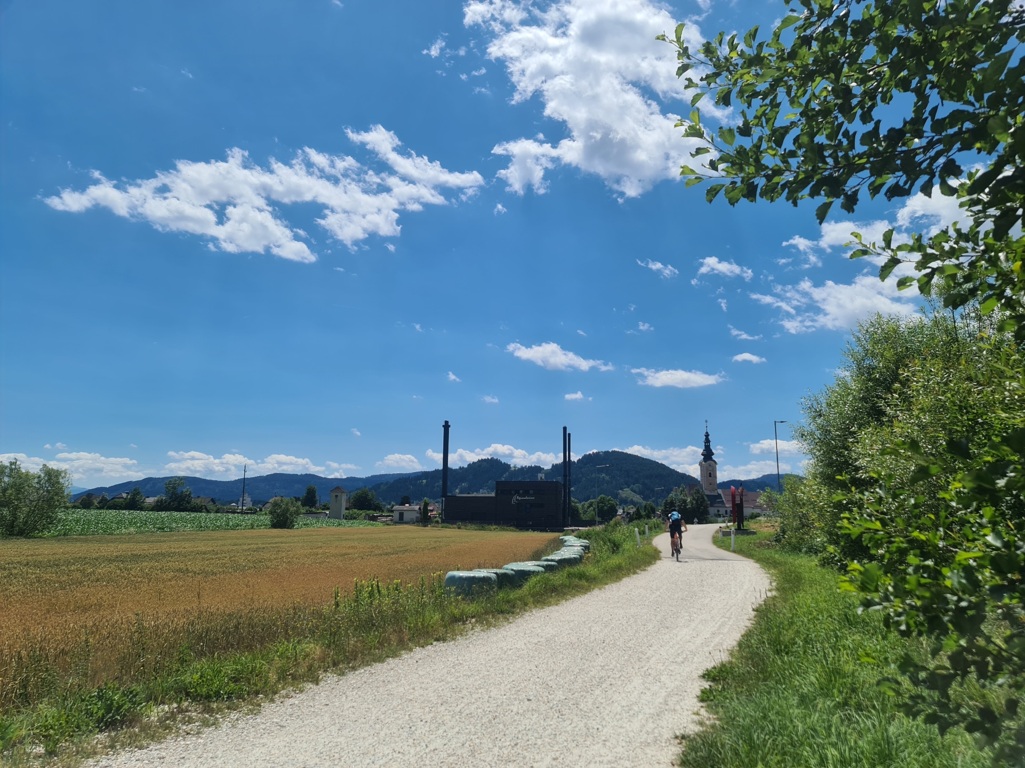The image shows a rural scene in Feldkirchen in Kärnten, Austria, under a bright blue sky with scattered white clouds. A person is cycling on a gravel path that curves to the right. To the left of the path, a field of golden grain is visible, bordered by green vegetation and some hay bales wrapped in green plastic. In the background, there is a cluster of buildings, including a church with a tall steeple, and a dark industrial-looking structure with two tall smokestacks. Rolling green hills form the backdrop. Lush green trees and foliage are present on the right side of the path and in the foreground.